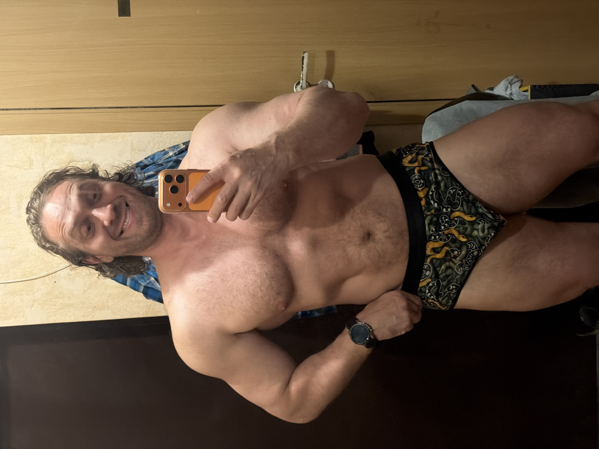 An indoor mirror selfie taken in Saint Petersburg, Russia, features a smiling man with a muscular physique. The man, appearing to be in his 30s or 40s, has long, wavy dark hair and a beard. He is captured from the waist up, holding an orange-cased smartphone with a triple camera setup in his right hand, which is angled towards himself to take the photo.
He is shirtless, showcasing his well-defined chest and abdominal muscles, and is wearing patterned boxer briefs that are dark green/black with yellow/gold, swirling snake-like or abstract designs. A dark-faced smartwatch with a black strap is visible on his left wrist. A blue plaid shirt is draped over his left shoulder and arm.
The background primarily consists of light brown wooden paneling, possibly a wardrobe or wall. In the lower left, a dark, reflective surface, possibly a television or a piece of furniture, is visible. Behind his right hip, a white towel or cloth and other indistinct objects are present. The man is looking directly at the camera with a wide, confident smile. No visible text is present in the image.