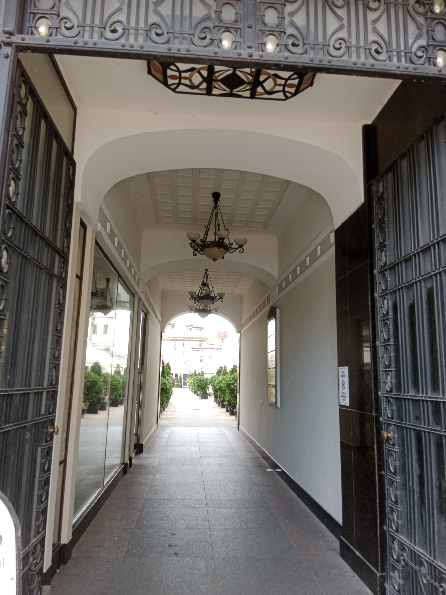 The image depicts a stone-paved walkway leading into a building, framed by open metal gates on either side. The walkway is lined with potted green plants and features decorative ceiling lights. The architecture suggests an older, possibly European style. The location is identified as Bydgoszcz, Poland. No people or animals are visible in the frame. The lighting is natural, casting soft shadows, indicating it is daytime. The scene is static and appears to be an entrance to a commercial or residential building.