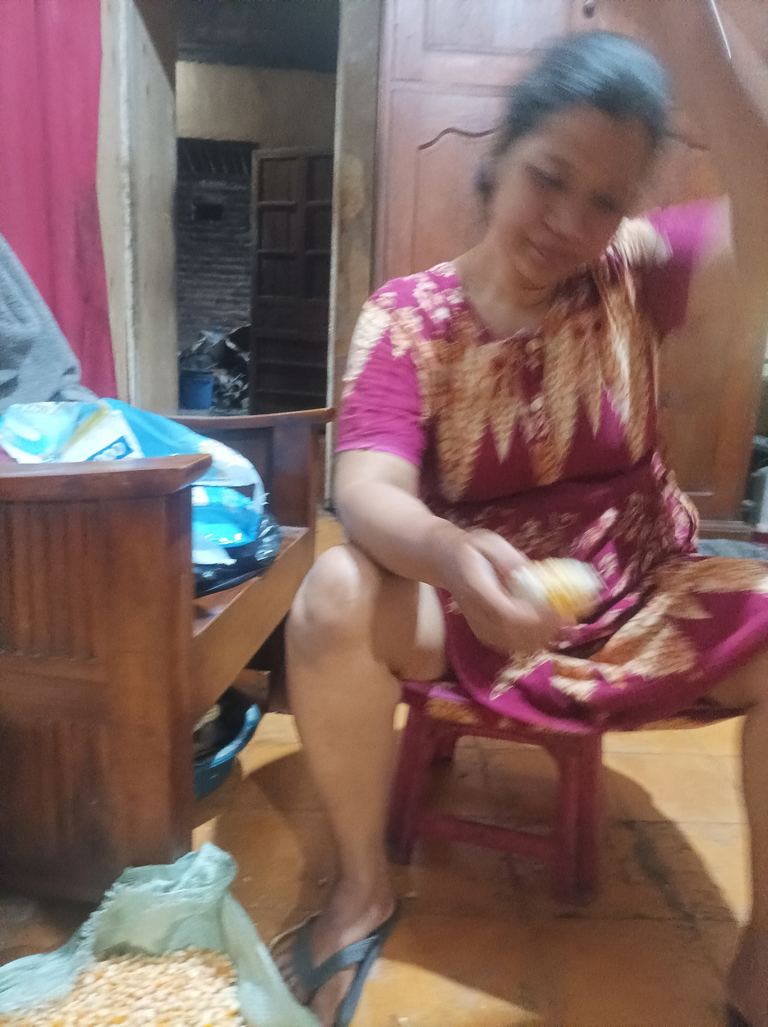 An out-of-focus, full-shot image shows a woman sitting on a small red stool in what appears to be a modest indoor dwelling, possibly in Canga'an, Indonesia. She is wearing a maroon and gold patterned dress and her right hand is reaching towards her face while holding a small, light-colored object. Her left leg is bent, and her right leg is extended. To her left, a wooden chair with a blue bag on it is visible. In the foreground, a light blue bag filled with what appears to be corn kernels is on the floor, and the woman's feet, wearing dark flip-flops, are visible near it. The background is cluttered with wooden furniture and what looks like a doorway leading to another room. The lighting is dim, suggesting an indoor setting during the day or evening.