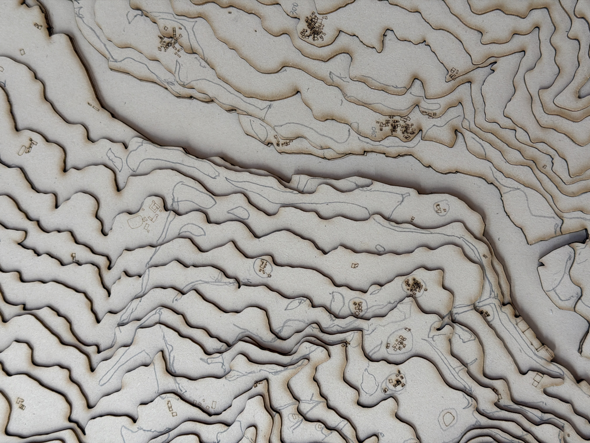 The image shows a topographical map model, likely made of laser-cut wood or cardboard. The model depicts a rugged terrain with contour lines indicating elevation changes. Scattered across the surface are small, abstract representations of buildings or structures, some clustered together to suggest a village or settlement. Faint lines, possibly indicating roads or paths, are also visible. The overall aesthetic is neutral and monochromatic, focusing on form and texture. The question about the location in Piacenza, Italy, cannot be answered from the visual information provided, as the image does not contain any identifying landmarks or textual clues related to a specific place.