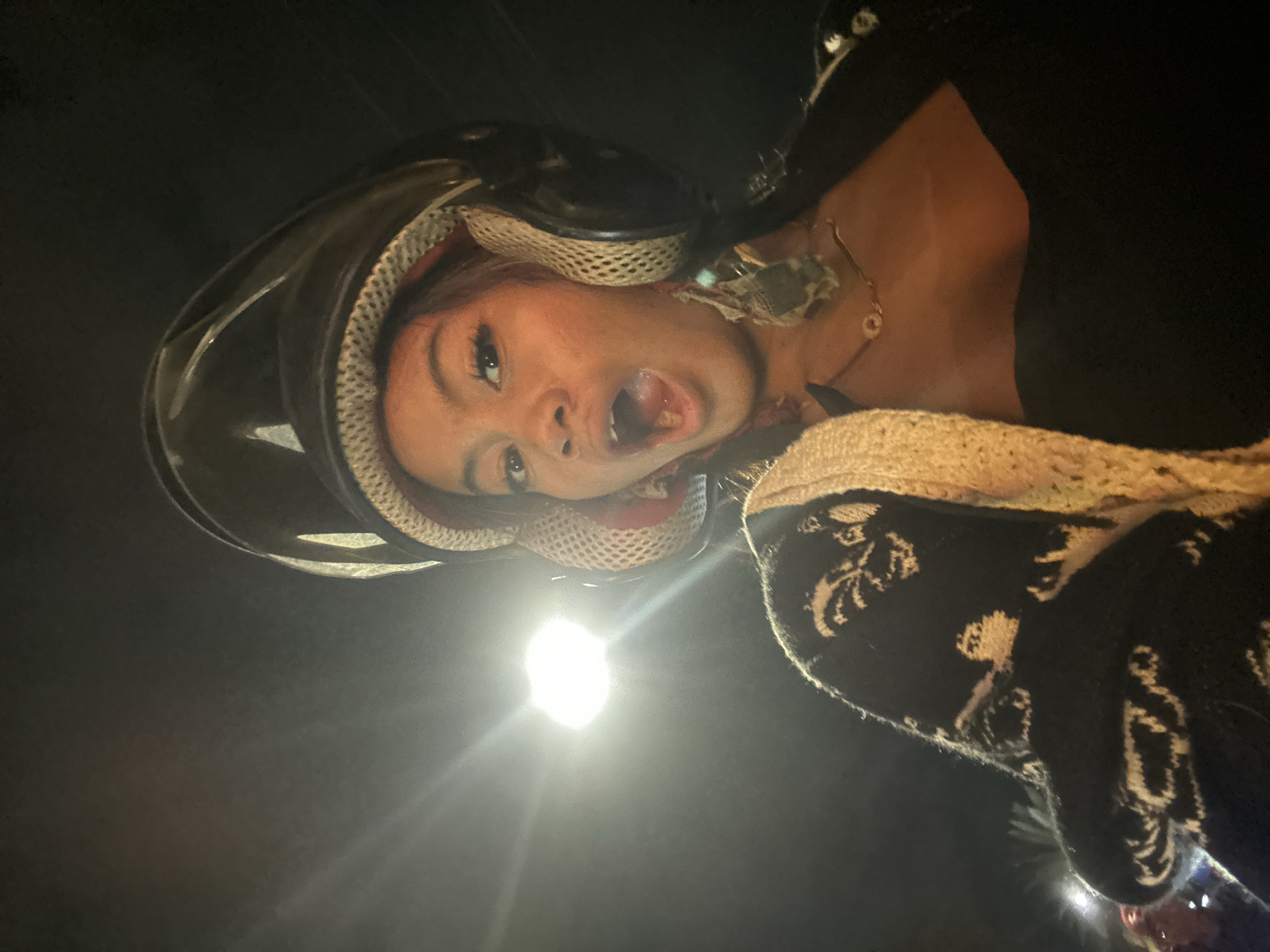 A woman is visible from a low angle, wearing a black helmet with a mesh interior. She has her mouth open in an expressive gesture. She is wearing a black top and a necklace with a circular pendant. Part of a black and beige patterned sweater or jacket is visible. The scene appears to be at night, illuminated by a bright light source which creates lens flare and light trails. Without additional information, it is impossible to confirm that the image was taken in North Jakarta, Indonesia.