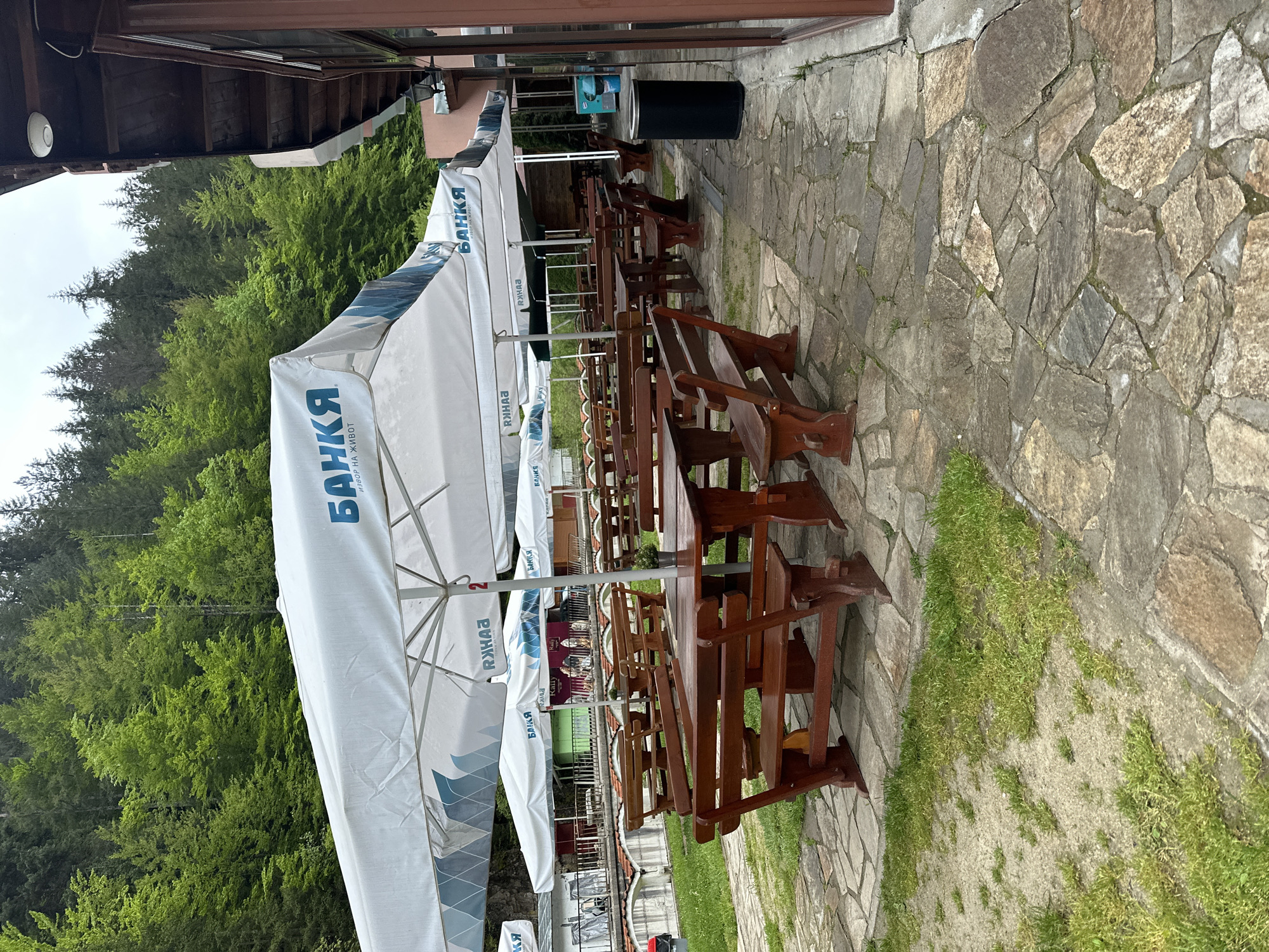 The image shows an outdoor seating area, likely a restaurant or café, situated adjacent to a building and overlooking a dense forest. Several rows of wooden picnic-style tables and benches are arranged on a stone-paved area. Large white umbrellas with blue lettering, possibly advertising a brand named "БАНКА" (Bank), provide shade over the tables. A black trash can is visible near the building. The environment suggests a rural or natural setting, with the forest as a prominent backdrop. The location of the scene is in Iagodina, Bulgaria.