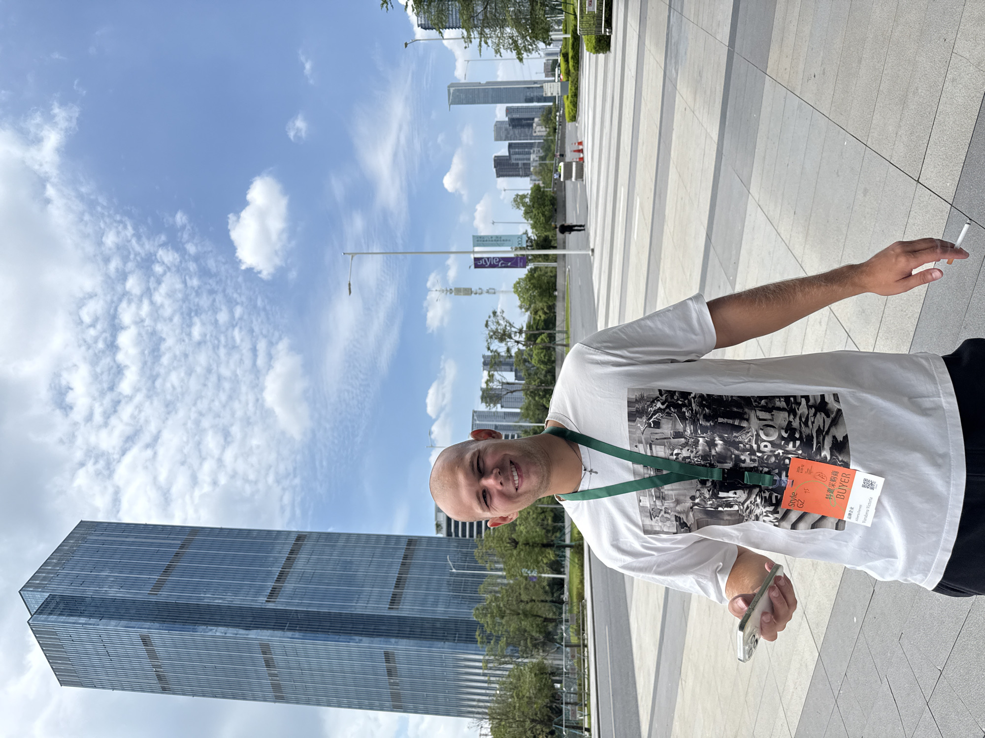 A bald, Caucasian male stands in an outdoor plaza with a skyscraper in the background, in Haizhu District, China. He is wearing a white t-shirt with a black and white graphic print, a green lanyard with a name tag, and black pants. The name tag reads "Style G2", "BUYER", and has Chinese characters. He is holding a smartphone in his left hand and a lit cigarette in his right hand. The plaza is paved with large gray tiles and has some greenery and streetlights visible. The sky is partly cloudy and blue. Other buildings and trees are visible in the distance.