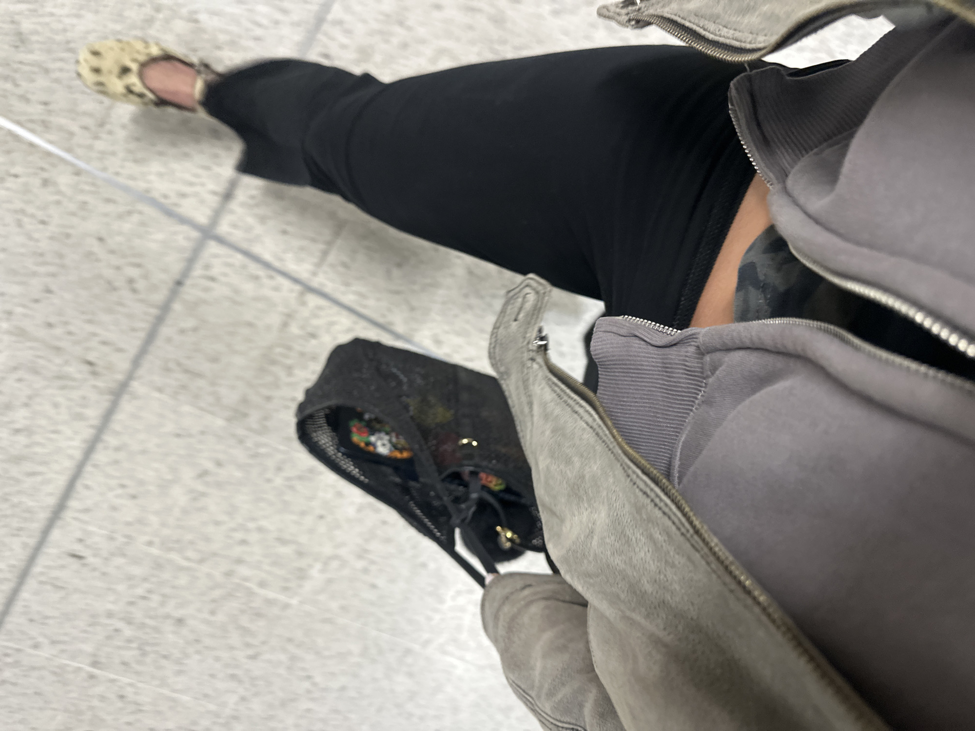 A single individual is depicted from a high-angle perspective, focusing on their lower torso and legs within an indoor public space in London, United Kingdom. The person is wearing a grey textured jacket, possibly suede, over a grey ribbed zippered hoodie. Below the waist, they wear black fitted trousers or leggings. A light-colored, flat shoe with a mottled pattern, resembling leopard print, is visible on the right foot. A black mesh drawstring bag, containing colorful items and golden metallic accents, is positioned near the left hip. The environment features a light grey tiled floor with visible grout lines. The specific location within London is not identifiable from the image, but the clean, utilitarian tiling is consistent with public transport hubs or commercial venues.