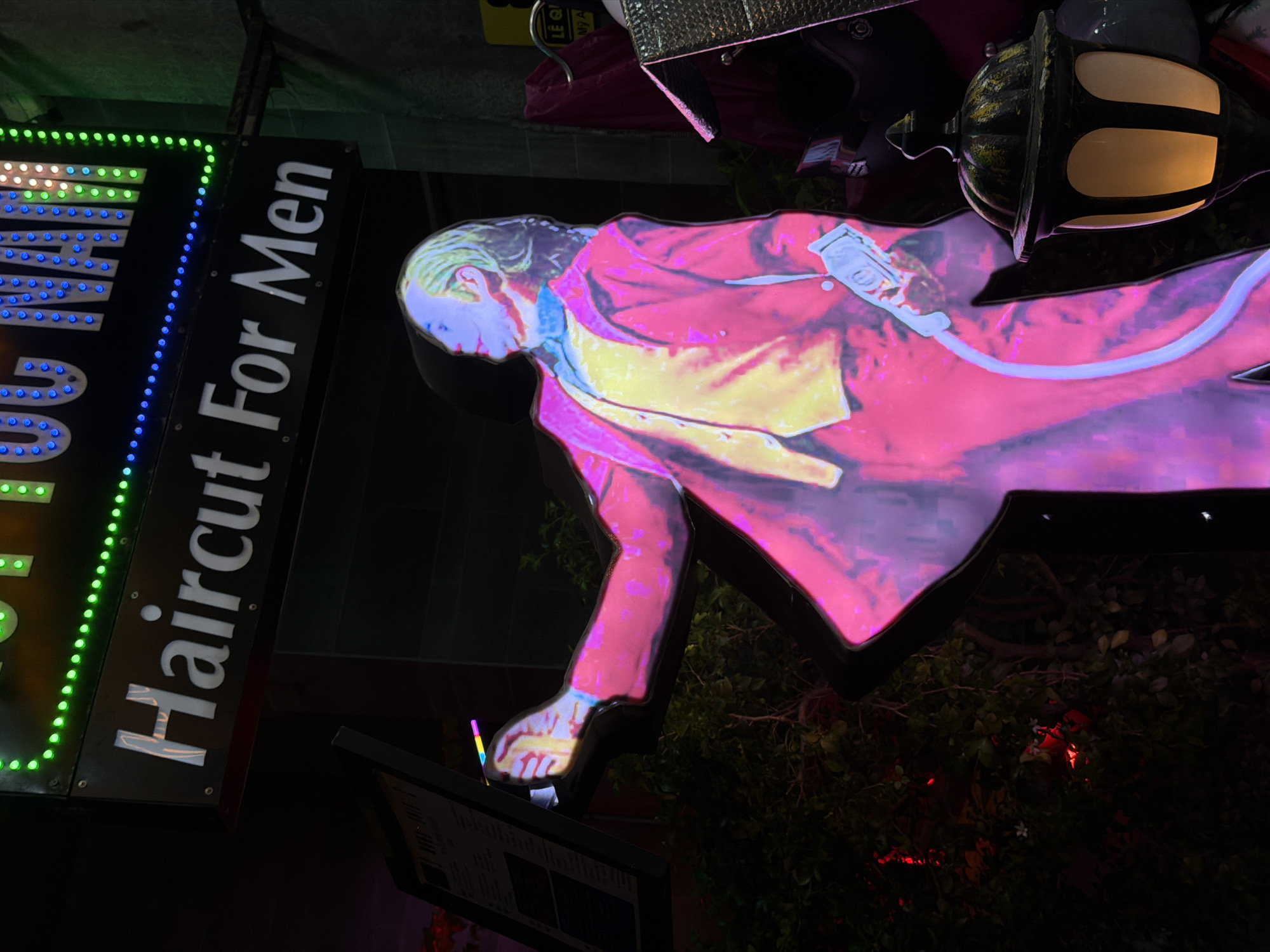 The image depicts a nighttime scene in Đà Nẵng, Vietnam, showcasing the signage for a "Haircut For Men" establishment. A rectangular sign with green and blue LED lights illuminates the phrase "Haircut For Men" in white lettering. Adjacent to the sign, a neon silhouette of a man in a suit holding a hair clipper is prominently displayed. The neon figure is rendered in shades of pink, yellow, and blue. A vintage-style lamppost with lit panels is positioned near the neon figure. A menu board stands underneath the neon figure and signage. Bushes and plants can be seen behind the signs and menu board.