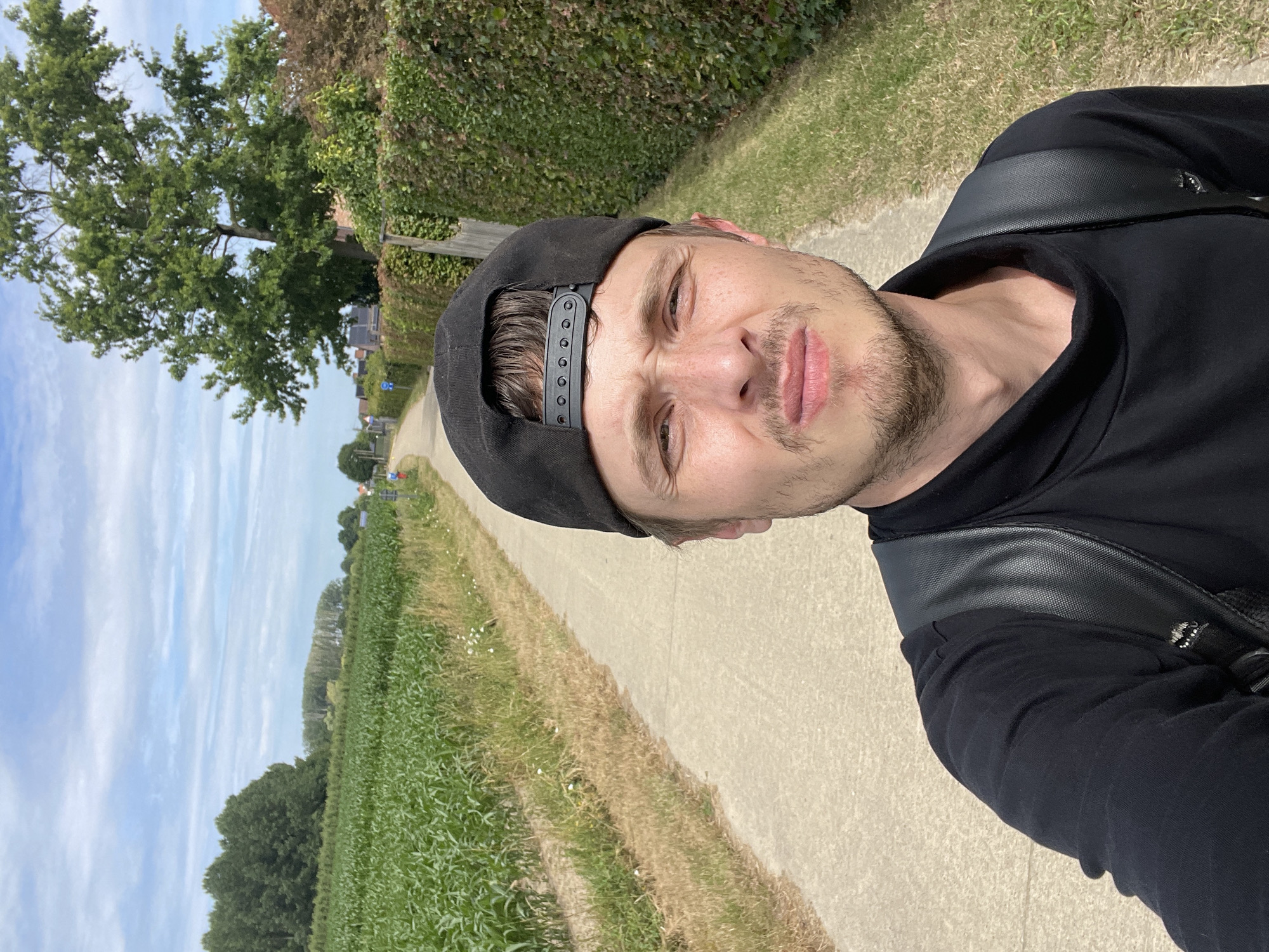 A man wearing a black baseball cap and black long-sleeved shirt is taking a selfie. He is standing on a paved path with a field of green crops on his left and a hedge and houses on his right. The path appears to be in a rural area, possibly on the outskirts of Diepenbeek, Belgium. The sky is blue with scattered clouds. The man's facial expression is ambiguous.