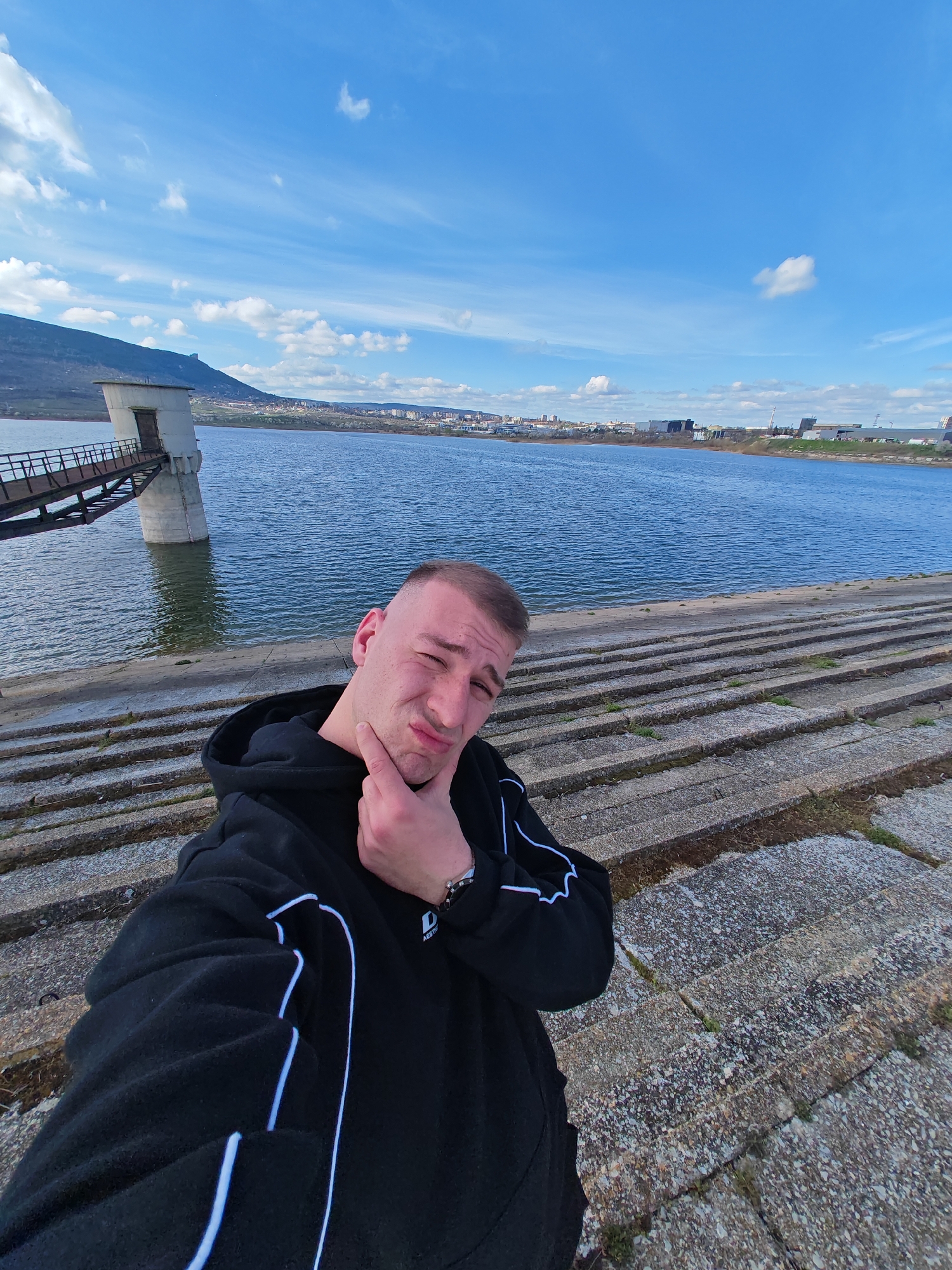 A fair-skinned male with a shaved head, wearing a black hoodie with white trim on the sleeves, is posing for a selfie outdoors. His left hand is raised to his chin, as though in contemplation or a playful expression of doubt. The background features a large body of water with a stone tower extending into it, connected by a metal walkway. A mountain is visible in the distance. The foreground shows the man standing on a series of wide, concrete steps leading down to the water's edge. The sky is blue with scattered white clouds. The location is identified as Unknown, Unknown.