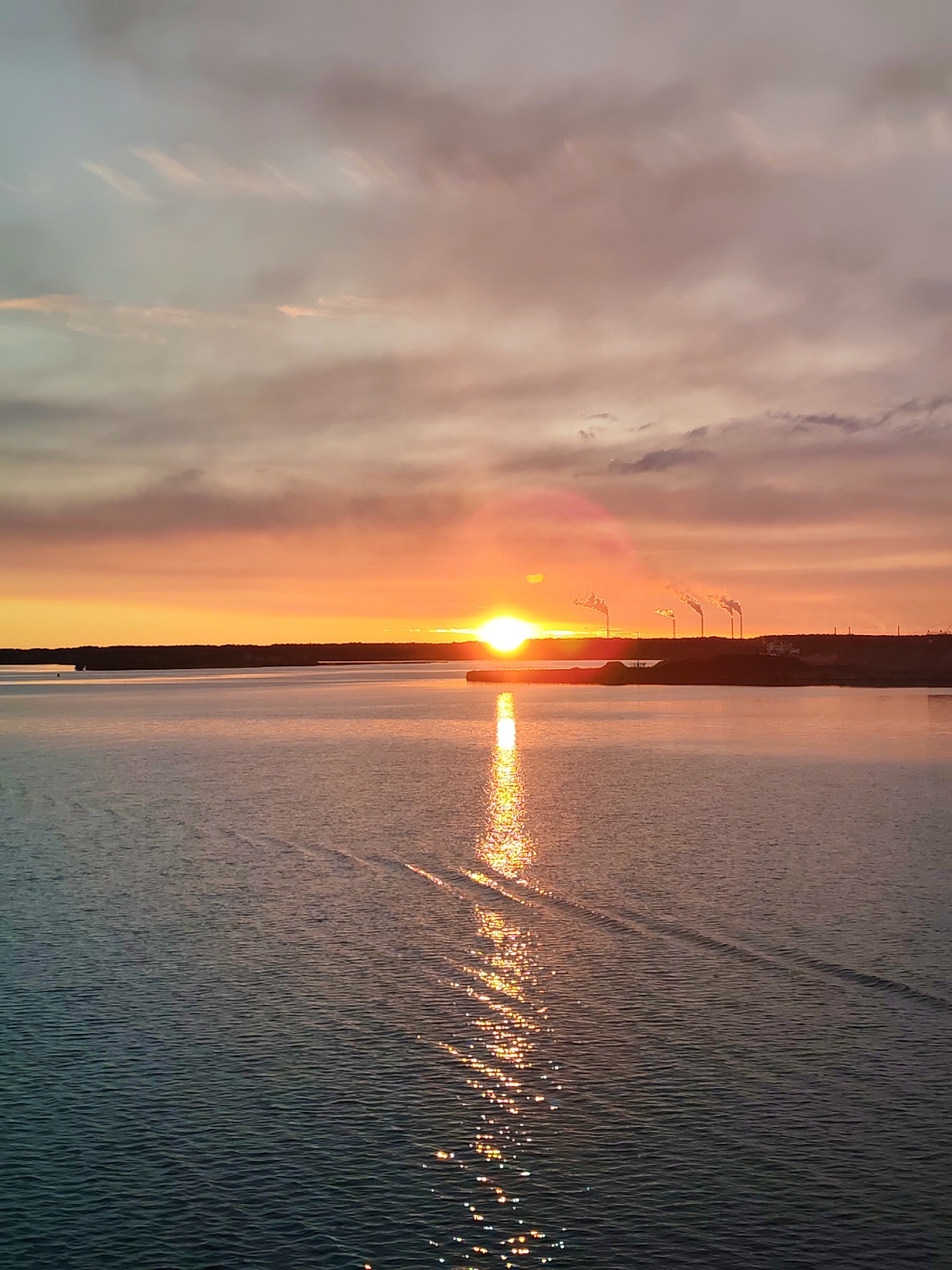 The image captures a sunset over a body of water, likely a lake or the sea. The sun, positioned near the horizon, reflects intensely on the water's surface, creating a shimmering effect. In the background, a dark shoreline is visible, punctuated by what appear to be industrial structures with smokestacks emitting plumes. The sky transitions from vibrant orange near the horizon to softer hues of pink and gray higher up. Ripples and wake patterns are visible in the water, suggesting the presence of a moving vessel. The location is unknown.
