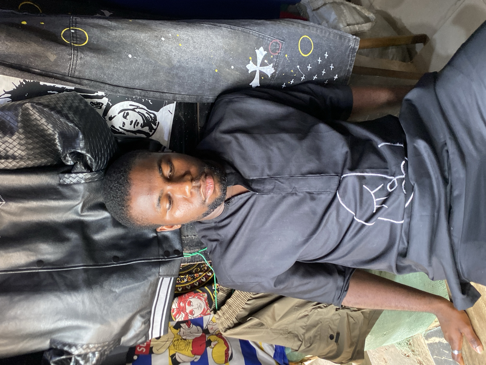 An overhead portrait-style shot, rotated 90 degrees clockwise, shows a young man resting or sleeping in what appears to be an indoor setting in Bukuru, Nigeria. His eyes are closed, and his mouth is slightly ajar, indicating a state of repose. He has short dark hair and a light beard.
The man is wearing a black, long-sleeved shirt featuring white, abstract, minimalist line art on the front, possibly depicting stylized faces or symbols. Underneath his arm and near his head, a black faux leather garment with a textured, possibly quilted, surface is visible. His right arm is extended, with a ring visible on his finger, resting on a light green woven mat or rug.
He is surrounded by various articles of clothing and textiles. A pair of dark grey or black distressed jeans with painted white splatters, yellow circles, white crosses, and a larger white cross-like symbol is laid out above his head. Below his left arm, several patterned fabrics are stacked: one with black and white comic-book style characters, another with red and yellow designs including a face resembling Medusa, and a partially visible "SUPREME" logo. Khaki-colored pants or shorts are also visible near his lower body.
The overall setting appears casual and possibly cluttered, suggestive of a personal space, a clothing store, or a market stall. The lighting is moderate, creating a relaxed atmosphere. A wooden furniture leg is partially visible in the background, further indicating an indoor environment.