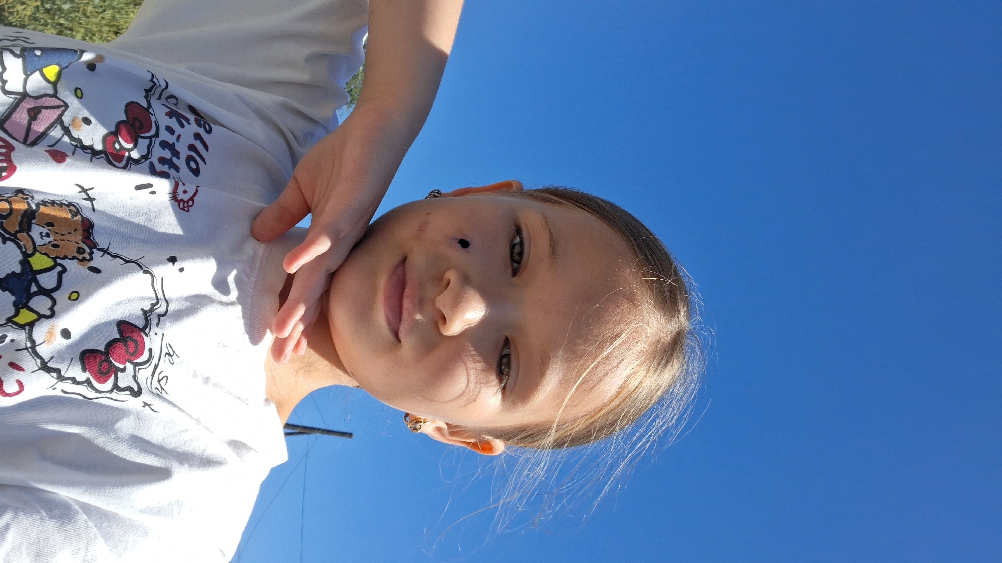 The media file is a close-up, portrait-oriented photograph of a young girl against a bright blue sky. The girl is the primary subject, and her face is angled towards the camera. She has light brown hair that appears windswept. Her eyes are a light brown or hazel color, and she has a slight smile. There are a few small marks on her cheek and nose. She is wearing a white t-shirt adorned with a graphic print featuring Hello Kitty characters. The t-shirt has a cartoon bear and red bows visible. Her left arm is extended, and her hand is resting near her face and on the t-shirt. The background is dominated by a clear, vibrant blue sky, suggesting it is daytime. The lighting indicates sunlight, casting shadows and highlighting the girl's features. The image appears to have been taken outdoors, possibly in a park or open area, given the hint of greenery at the top left corner of the original image. The overall mood of the photograph is casual and perhaps playful. There is no discernible text in the visible portion of the image other than the graphics on the t-shirt.