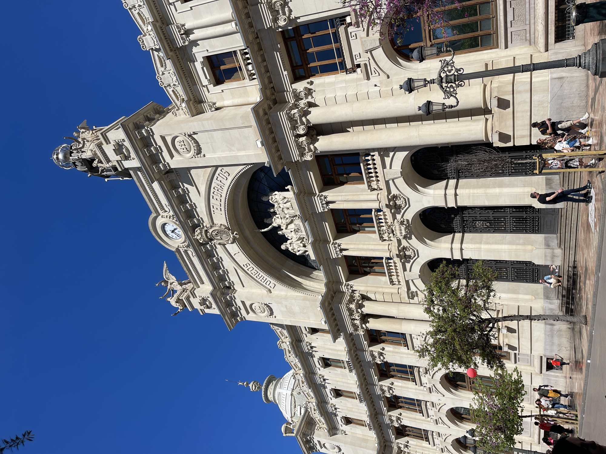 The image depicts the Correos building (Post Office) in Valencia, Spain, from a low-angle perspective. The building, made of light-colored stone, features ornate details, including columns, arches, decorative sculptures, and inscriptions that read "CORREOS TELEGRAFOS." A clock is visible on one of the building's sections. The sky is a clear, deep blue. People are visible in front of the building on the sidewalk, including individuals walking and others sitting on steps. A street lamp is also visible near the people. A tree is positioned near the building's base.
