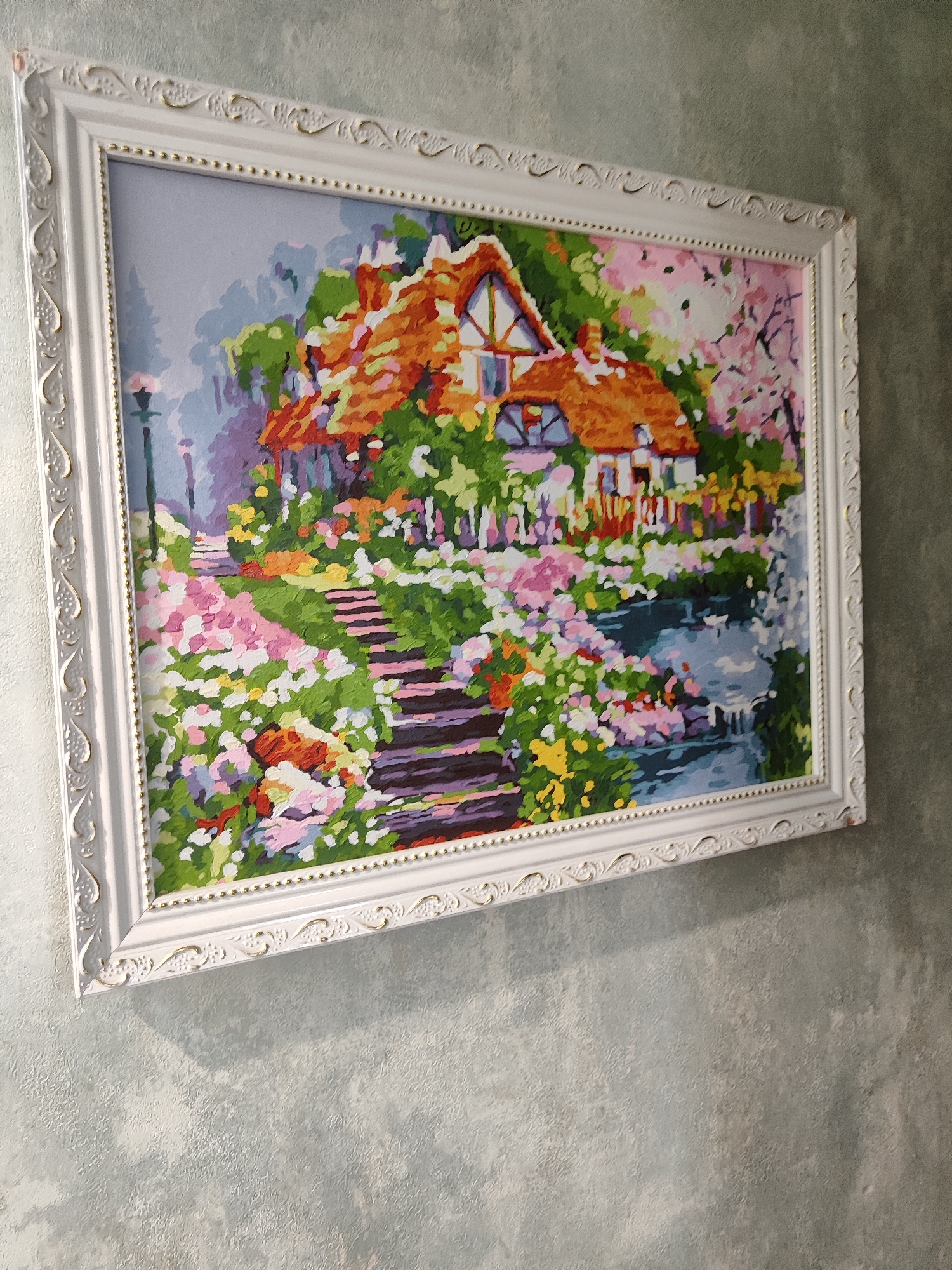 A framed painting of a cottage and garden is displayed on a textured wall. The painting depicts a rustic cottage with an orange roof, surrounded by abundant flowers in various shades of pink, white, and yellow. A stone pathway leads up to the cottage, and a body of water, possibly a pond or stream, is visible in the foreground. The frame is white with ornate detailing and a gold beaded inner edge. The wall appears to be a light grey with a subtle textured pattern. The lighting suggests an indoor setting, with shadows cast on the wall by the painting. The scene is static and does not show any activity or individuals. The location is not specified in the image, but it is described as being in "Unknown, Unknown."