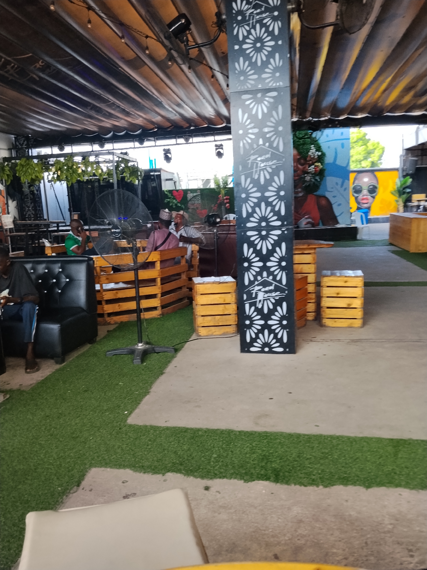 This image depicts an outdoor seating area at a venue, possibly a bar or restaurant, located in Howayi, Nigeria. A large, black, oscillating fan stands in the foreground, positioned on a patch of artificial turf. To the left, a person is seated on a black leather sofa. In the background, several people are seen sitting in wooden pallet-style seating arrangements. A tall, decorative pillar with white floral cutouts and a logo is prominently featured in the center of the frame. The ceiling overhead is constructed of corrugated metal panels with exposed lighting fixtures. The overall environment appears to be a casual, open-air gathering space.