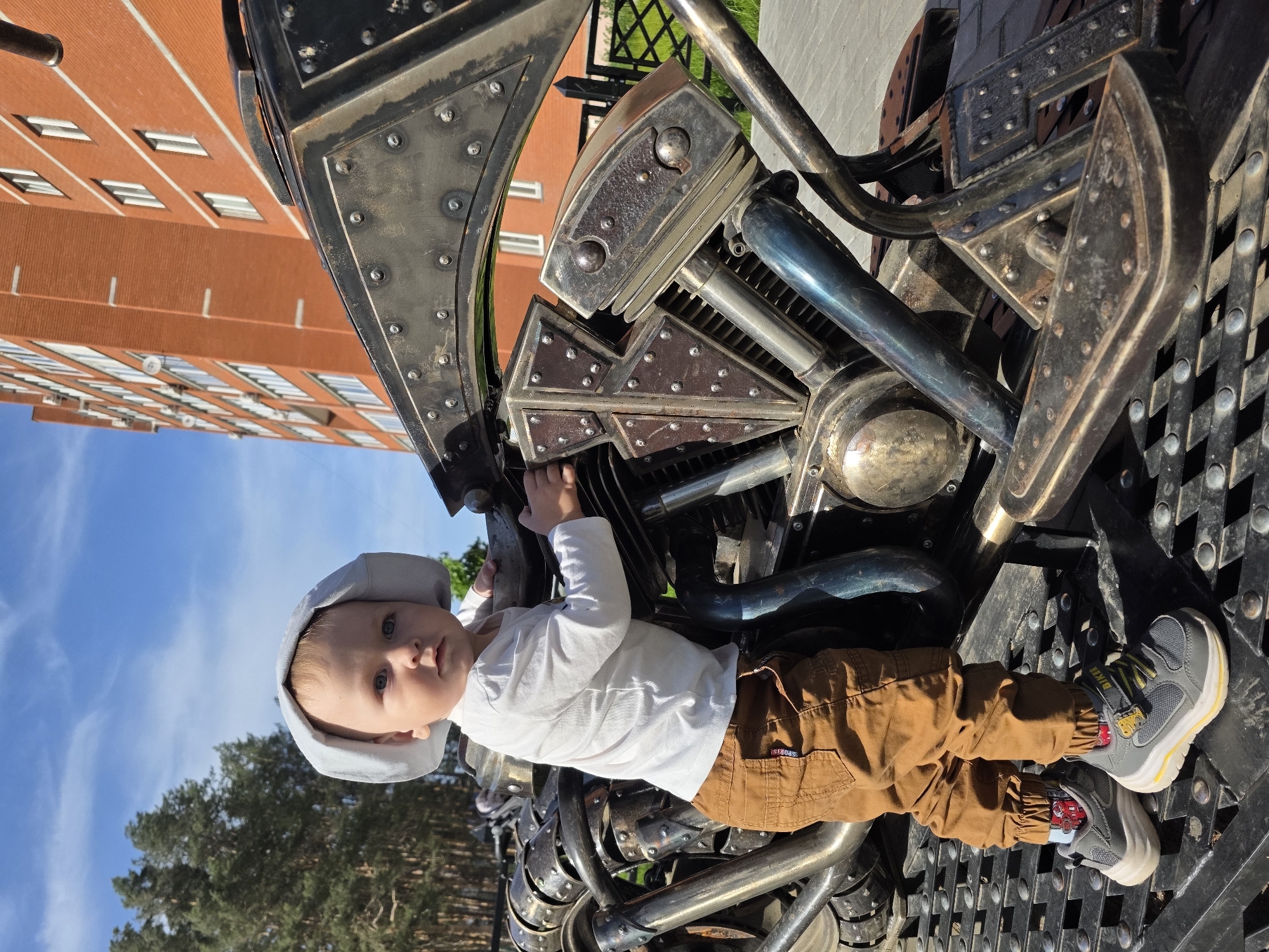 A young child sits on a metal sculpture of a motorcycle in what appears to be a public area, possibly a park or recreational space. The child wears a light grey hat, a white long-sleeved shirt, and brown pants with small shoes. The child is partially supported by the motorcycle’s frame. The motorcycle sculpture features a complex design of riveted plates, tubes, and other metal components. Behind the child and the motorcycle, there is a brick building with multiple windows, suggesting an apartment or office complex. Trees and a blue sky with clouds are visible in the background. The location appears to be the city of Chaikovsky, Russia.
