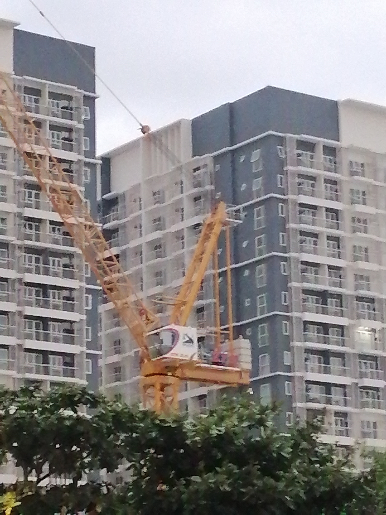 The image depicts a construction site in Davao City, Philippines. A large yellow tower crane is prominently featured, extending diagonally across the frame. Behind the crane, several multi-story buildings, possibly apartment complexes or condominiums, are under construction or recently completed. These buildings feature a modern architectural style with multiple windows and balconies. The sky is overcast, suggesting a cloudy or dull weather day. In the foreground, the tops of several lush green trees are visible, indicating a potentially urban environment with green spaces. The scene is consistent with ongoing urban development projects in a bustling city.