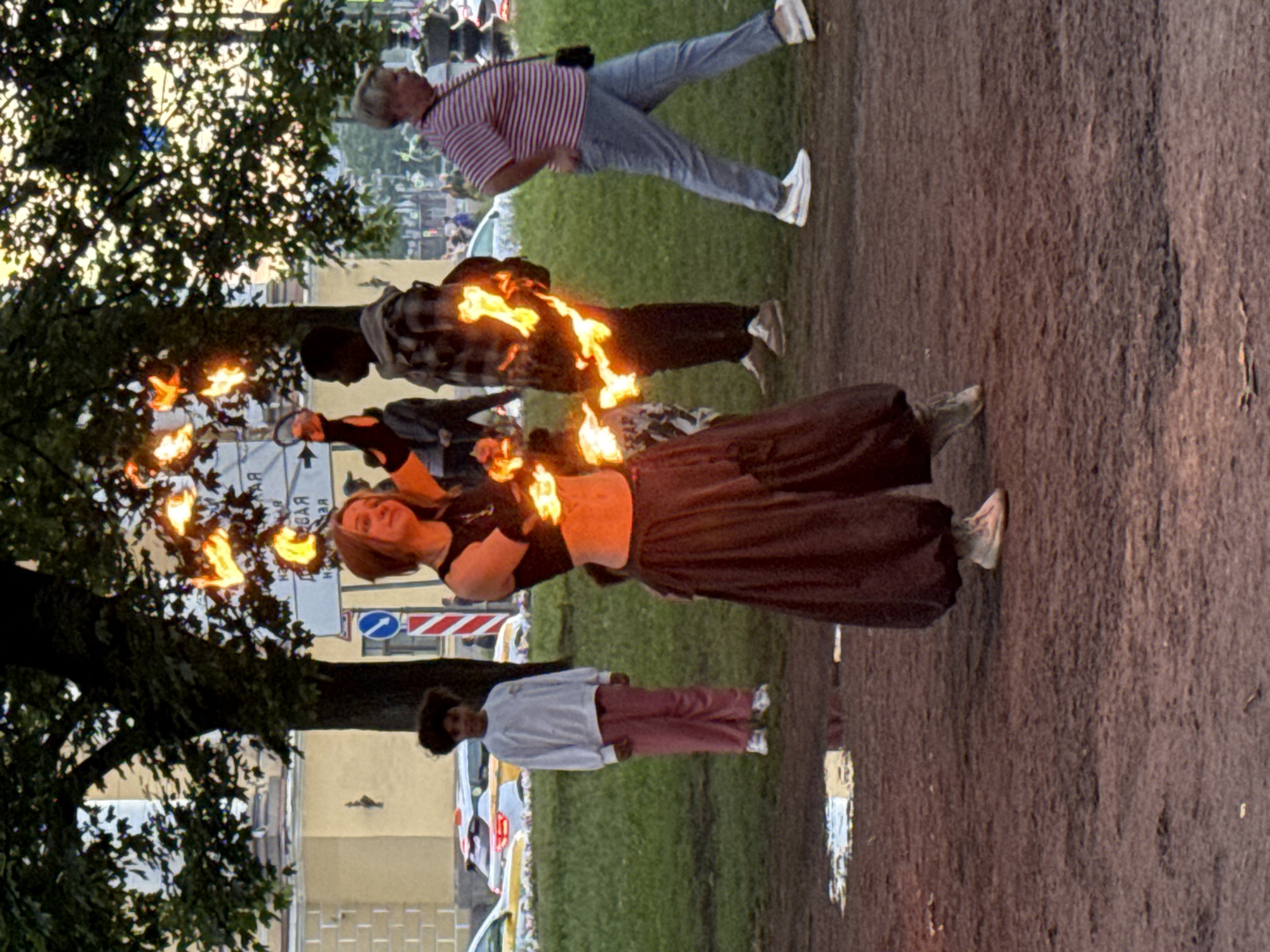 A young woman performs with fire in front of a crowd in St. Petersburg, Russia. She is wearing a black crop top and a long, dark skirt, with her midriff exposed, and she is holding a ring of fire. The fire is burning brightly and casting light on her and the people around her. In the background, other people are visible, including a child wearing a white sweatshirt and pink pants, and a man in a plaid shirt. There are cars and buildings in the distance, and trees line the street. The ground is covered in dirt and grass. The lighting suggests it is dusk or early evening.
