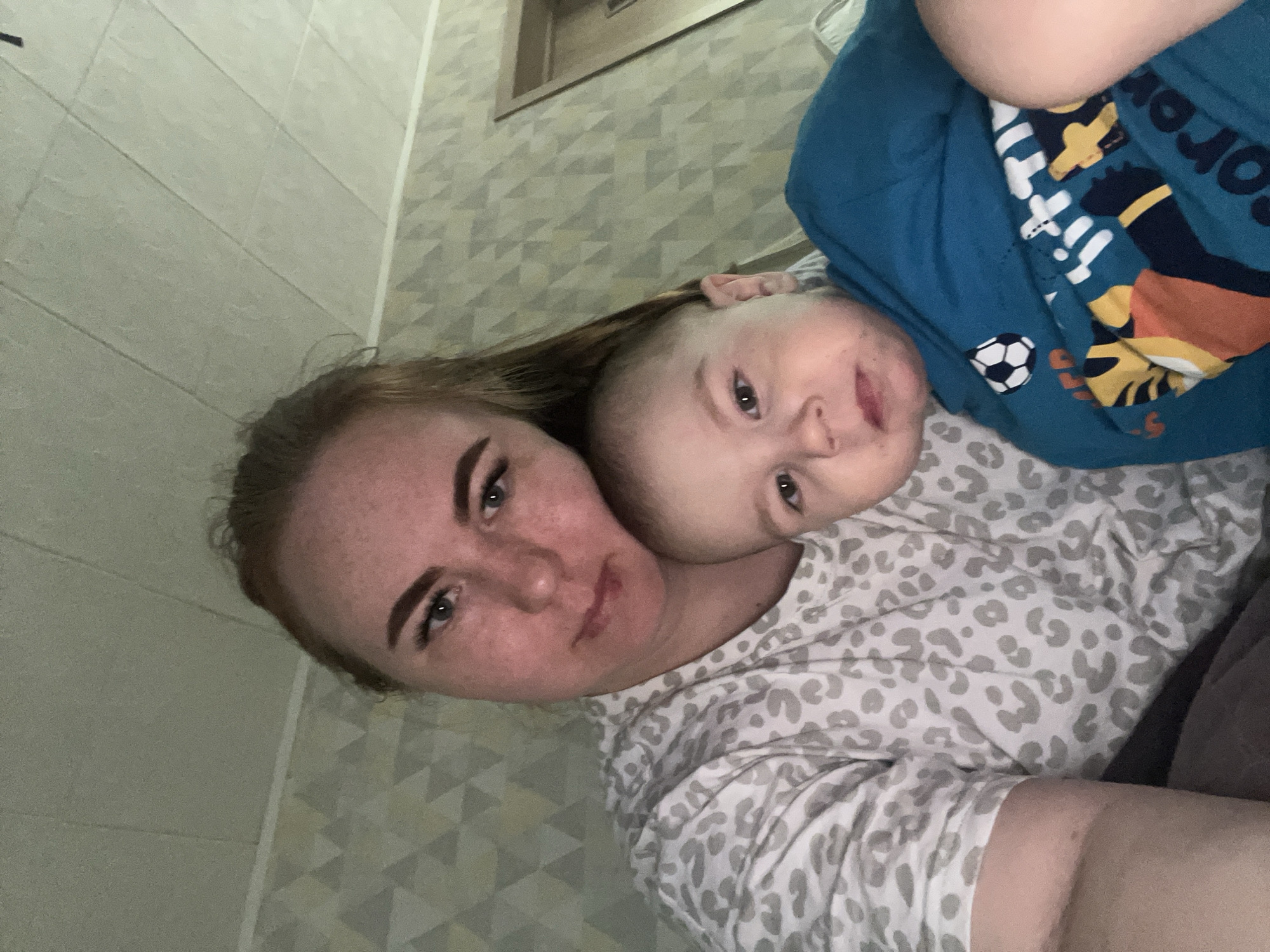 A woman with fair skin and freckles is holding a baby wearing a blue shirt with a soccer ball. They appear to be lying down together, the baby partially on the woman. The woman has reddish-blonde hair and is looking at the camera. The background shows a tiled wall and a patterned surface, possibly a bedspread or wall covering. There is no definitive indication that the location is in Chaikovsky, Russia.