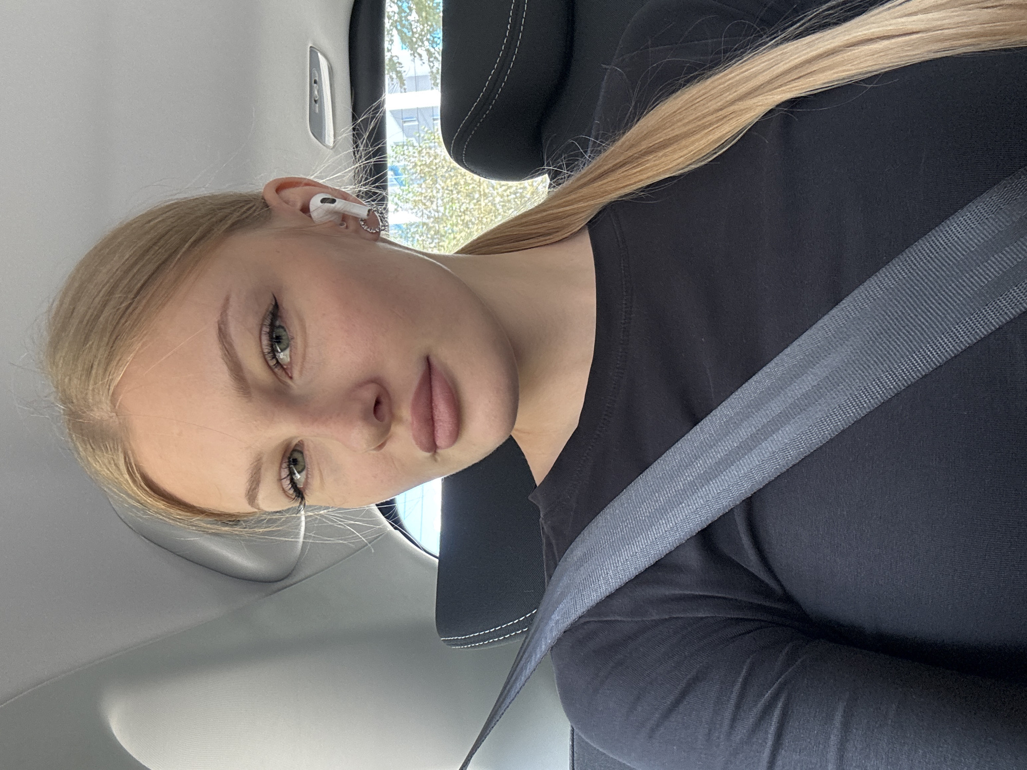 A young Caucasian woman with blonde hair pulled back in a ponytail, wearing a black long-sleeved shirt and a grey seatbelt, is seated in the back of a vehicle. She is wearing white wireless earbuds and a silver hoop earring on her left ear. She is looking towards the camera with a neutral expression. The interior of the car is visible, including the headrest of the seat in front of her and the car's ceiling. Through the window behind her, trees and a building are visible, suggesting an urban or suburban environment, possibly in the city of Perm, Russia. The lighting indicates it is daytime.