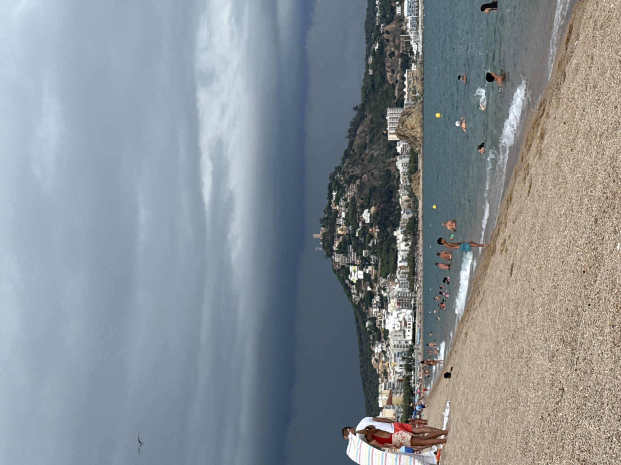The image captures a beach scene in Blanes, Spain, with a dramatic, dark, stormy sky looming overhead. In the foreground, a sandy beach is populated with a few people, some standing and others partially obscured by a colorful beach towel. The water is a vibrant blue-green, with several individuals swimming and playing. The background reveals a coastal town nestled on a hillside, with numerous buildings and what appears to be a castle or fortress at the summit. The overall atmosphere is one of impending weather change, with the contrast between the dark sky and the lively beach activities.