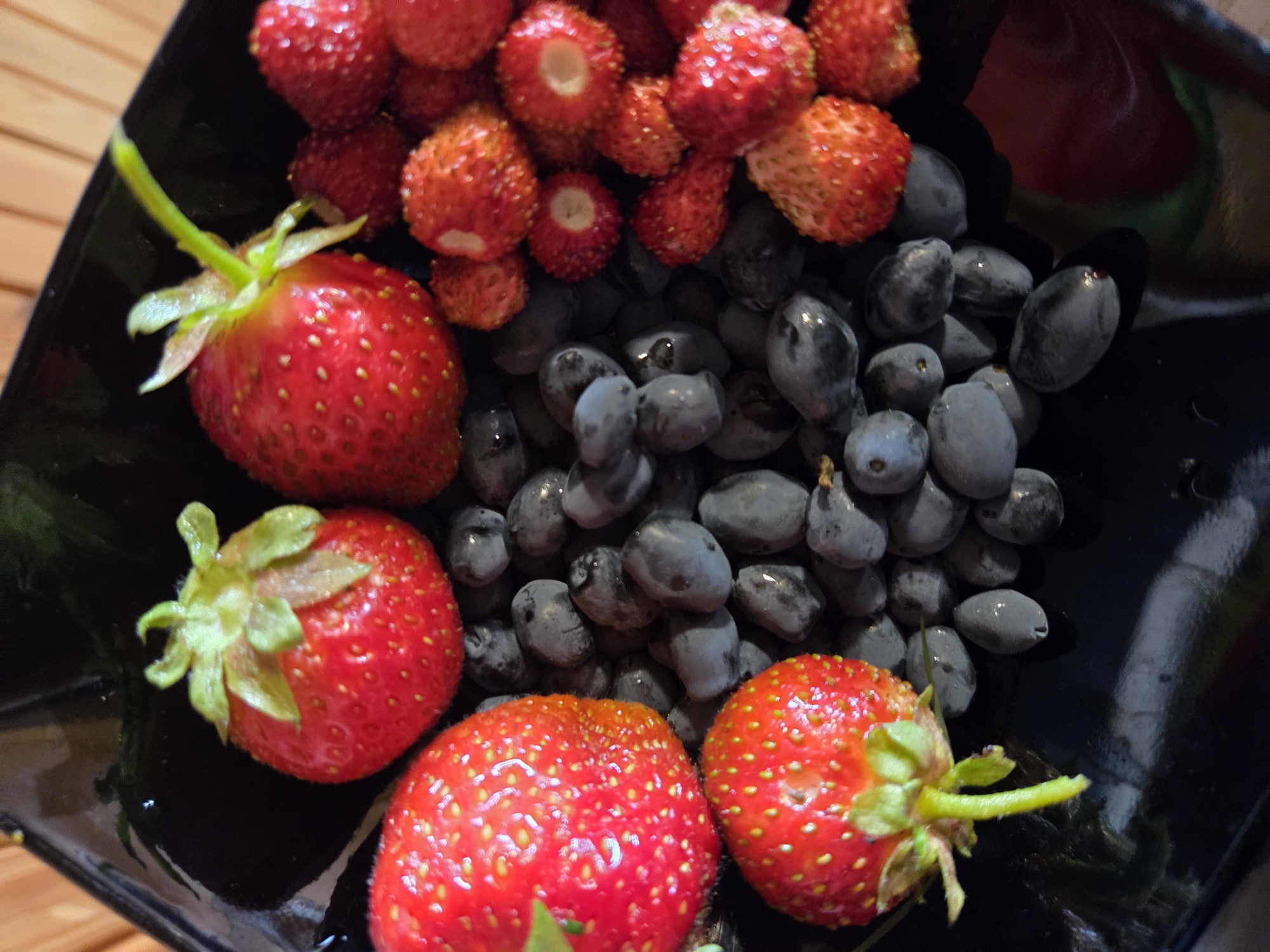 A close-up, overhead shot shows a black bowl filled with strawberries and blueberries. The strawberries are bright red and plump, with their green leaves and stems still attached. The blueberries are dark blue and round, scattered among the strawberries. The bowl itself is black and glossy, reflecting the light. The scene is set on a wooden surface, possibly a table or counter, with a faint wooden grain visible in the background. The lighting is bright and natural, suggesting daytime. The focus is sharp on the fruits, highlighting their textures and vibrant colors. There are no people visible, and no text is present in the image. The overall impression is one of freshness and abundance, perhaps a preparation for a meal or a simple fruit snack.