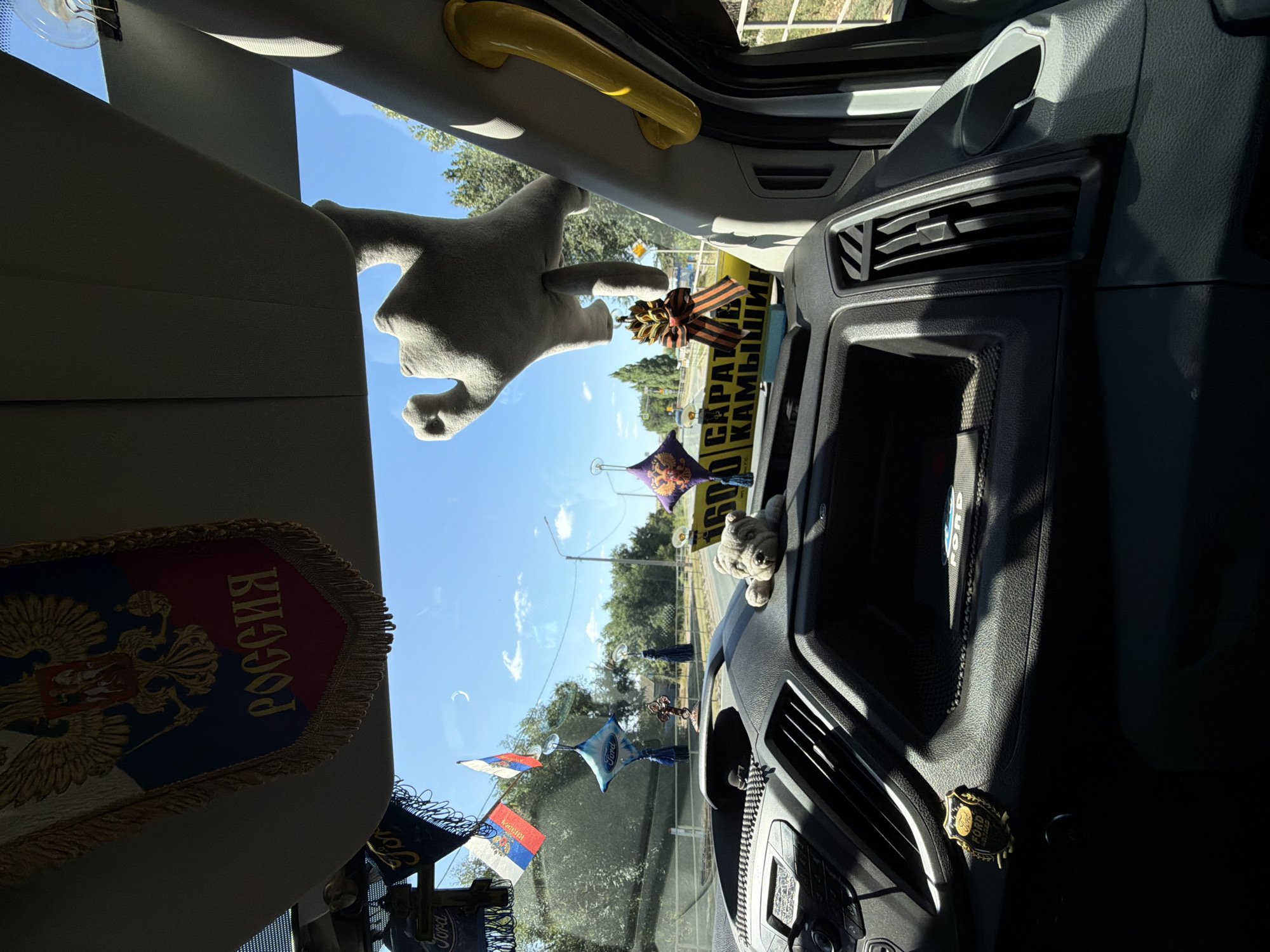 The image shows the interior of a vehicle looking out through the windshield on a sunny day. Inside the car, a banner with the Russian coat of arms and the word "РОССИЯ" (RUSSIA) is draped from the sun visor. Several small Russian flags are hanging from the windshield. A stuffed grey hand hangs from above, and a plush dog is placed on the dashboard. A yellow sign with Russian text is visible through the windshield. Outside, a road with trees and power lines can be seen. The location is identified as Оленье, Russia.