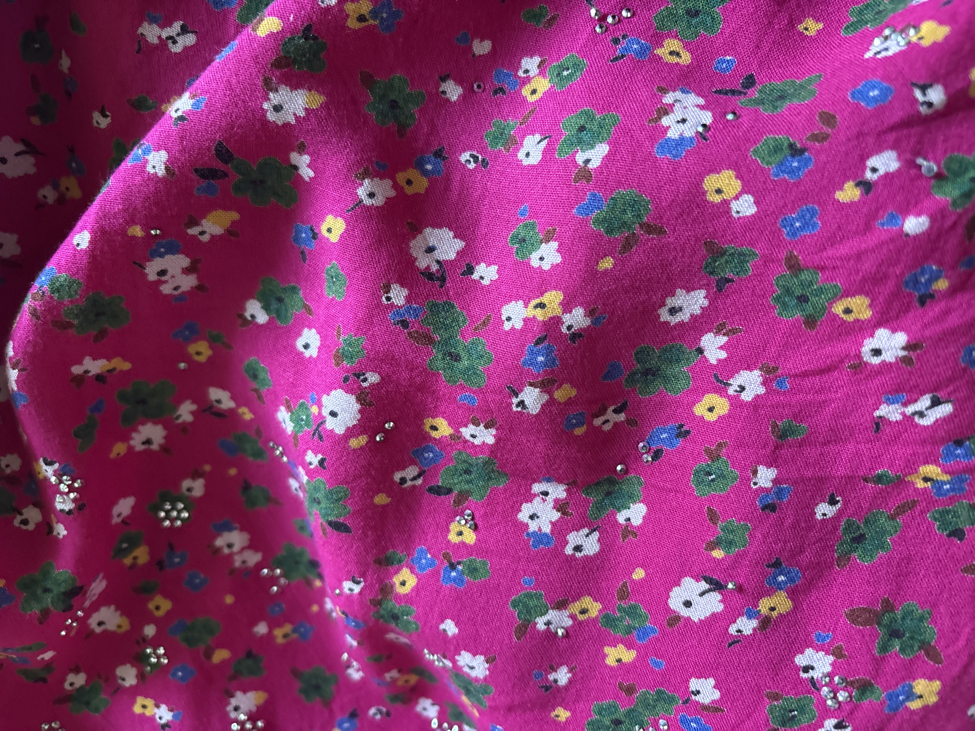 The image is a close-up, high-angle shot of a bright pink fabric with a floral pattern. The fabric is draped and creased, creating shadows and highlights that add depth to the image. The floral pattern consists of small flowers in various colors, including white, green, yellow, and blue, with some flowers having small, sparkling embellishments. The background is entirely the pink fabric. There is no visible information about the location in the city of Unknown, Unknown.
