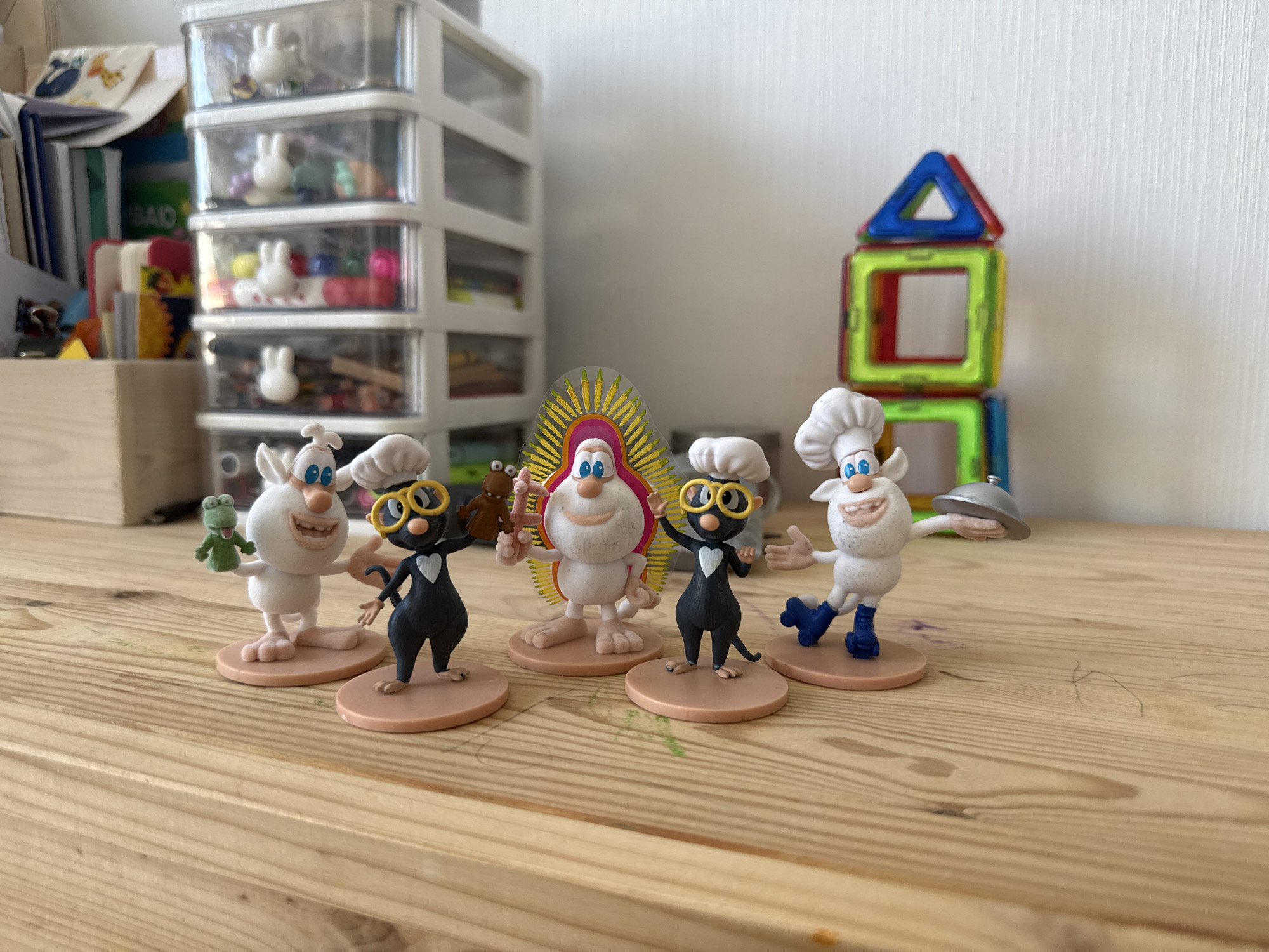 Five cartoon-like figurines are arranged on a wooden surface. From left to right, the first figurine is white with blue eyes, holding a green frog. The second figurine is black with yellow glasses and is wearing a white heart-shaped bib. The third figurine is white with blue eyes and a halo-like headdress. The fourth figurine is black with yellow glasses and a white heart-shaped bib, similar to the second. The fifth figurine is white with blue eyes, wearing a chef's hat and blue shoes, and holding a silver serving dome. In the background, there is a white plastic storage unit with multiple drawers filled with small items, and a colorful magnetic building set. The scene appears to be indoors, likely on a desk or shelf. The presence of these specific figurines suggests a connection to the animated series "The Three". This image does not provide any information about the city of Lyubertsy, Russia.