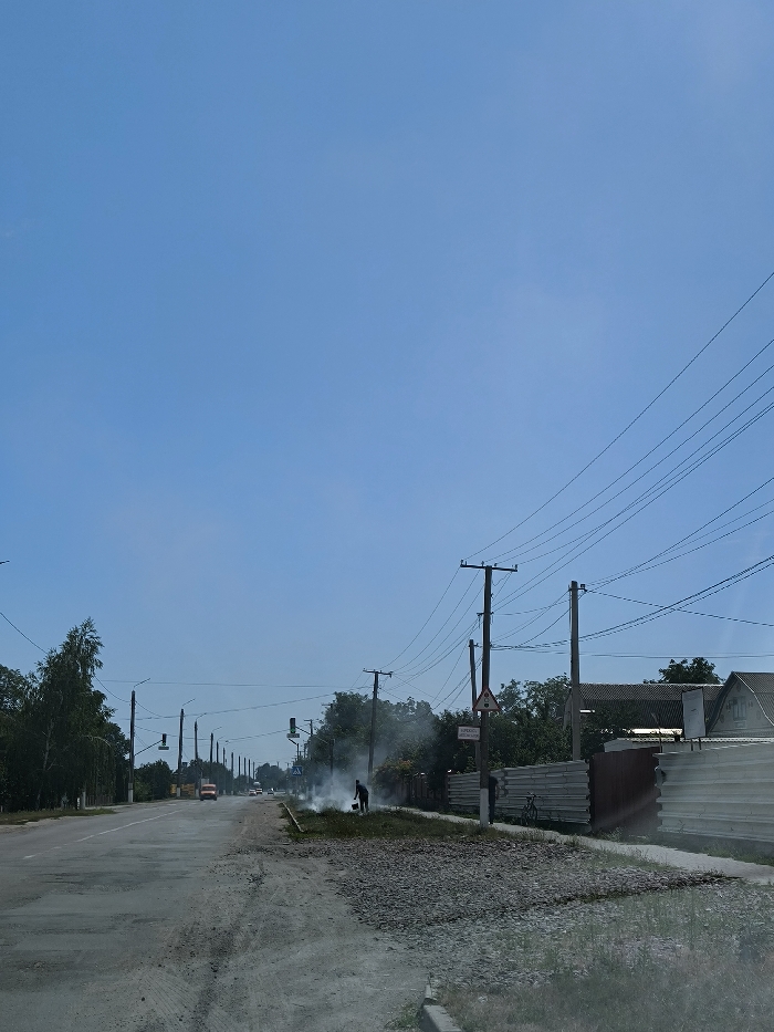 A clear blue sky dominates this daytime exterior image captured in Illintsi, Ukraine. The scene depicts a two-lane asphalt road with a worn surface and gravel shoulders extending into the distance. On the right shoulder, a lone individual, dressed in dark attire, operates a device that generates a substantial plume of white smoke. Adjacent to this activity, a large deposit of grey gravel covers part of the roadside. To the immediate right, a corrugated metal fence borders partially visible residential structures. A bicycle is parked against this fence. Numerous wooden utility poles with multiple overhead power lines are positioned along both sides of the road. A triangular warning sign and a rectangular white sign with the Cyrillic text "ОБЕРЕЖНО, ВИСОКА НАПРУГА" (CAUTION, HIGH VOLTAGE) are affixed to a utility pole on the right. Green trees and other vegetation line both road margins. In the mid-distance on the road, a light-colored truck and additional vehicles are visible, alongside traffic signals.