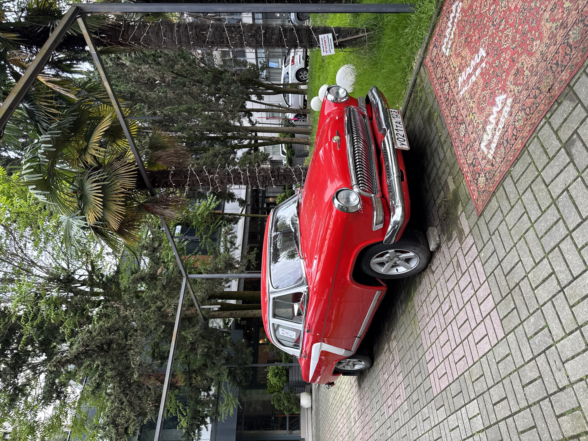 The image depicts a red vintage car parked on a brick pathway in Sochi, Russia. The car is a GAZ-21 Volga, recognizable by its distinctive grille and shape. It is parked adjacent to a strip of green grass and a red rug. In the background, there are trees and buildings, suggesting an urban environment. A metal frame structure is positioned near the car. The license plate reads Y021TA 123, identifying it as Russian. A sign is visible near the car, but the text is not clear. The scene appears to be a staged or preserved display, possibly for touristic purposes.