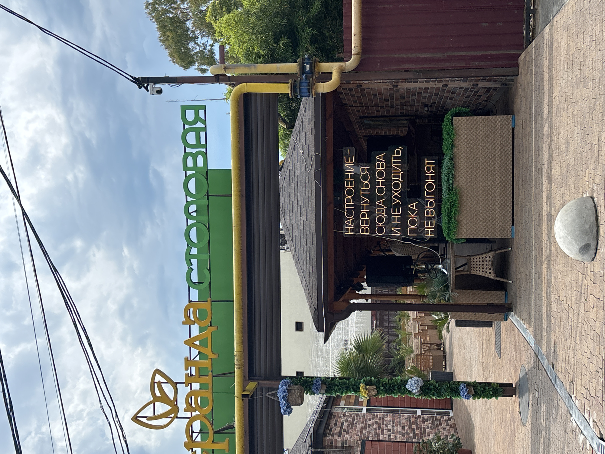 The image shows a street scene in Sochi, Russia, featuring a cafe or restaurant with a prominent sign and outdoor seating area. The sign, in yellow and green lettering, reads "Бранда столовая" (Brand Dining). The cafe has a covered seating area with tables and chairs, and decorative plants. A neon sign inside the seating area displays a humorous message in Russian. The overall atmosphere suggests a casual dining establishment in a warm climate.