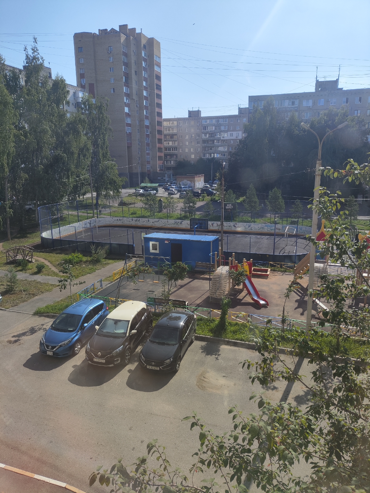 The image shows a residential courtyard in Ufa, Russia, under a clear blue sky. In the foreground, three parked cars are visible: a blue Nissan Qashqai, a black Renault Duster with a beige roof, and a black Ford Focus. Beyond the cars, a children's playground features a red slide, a sandbox, and swings. Adjacent to the playground is an enclosed sports court, likely for hockey or basketball, with a net at one end. In the background, several multi-story apartment buildings line the courtyard, with trees and vegetation interspersed throughout the scene. A blue portable structure, possibly a storage unit or changing room, is located near the sports court. The overall impression is of a typical urban residential area in Russia during daylight hours.