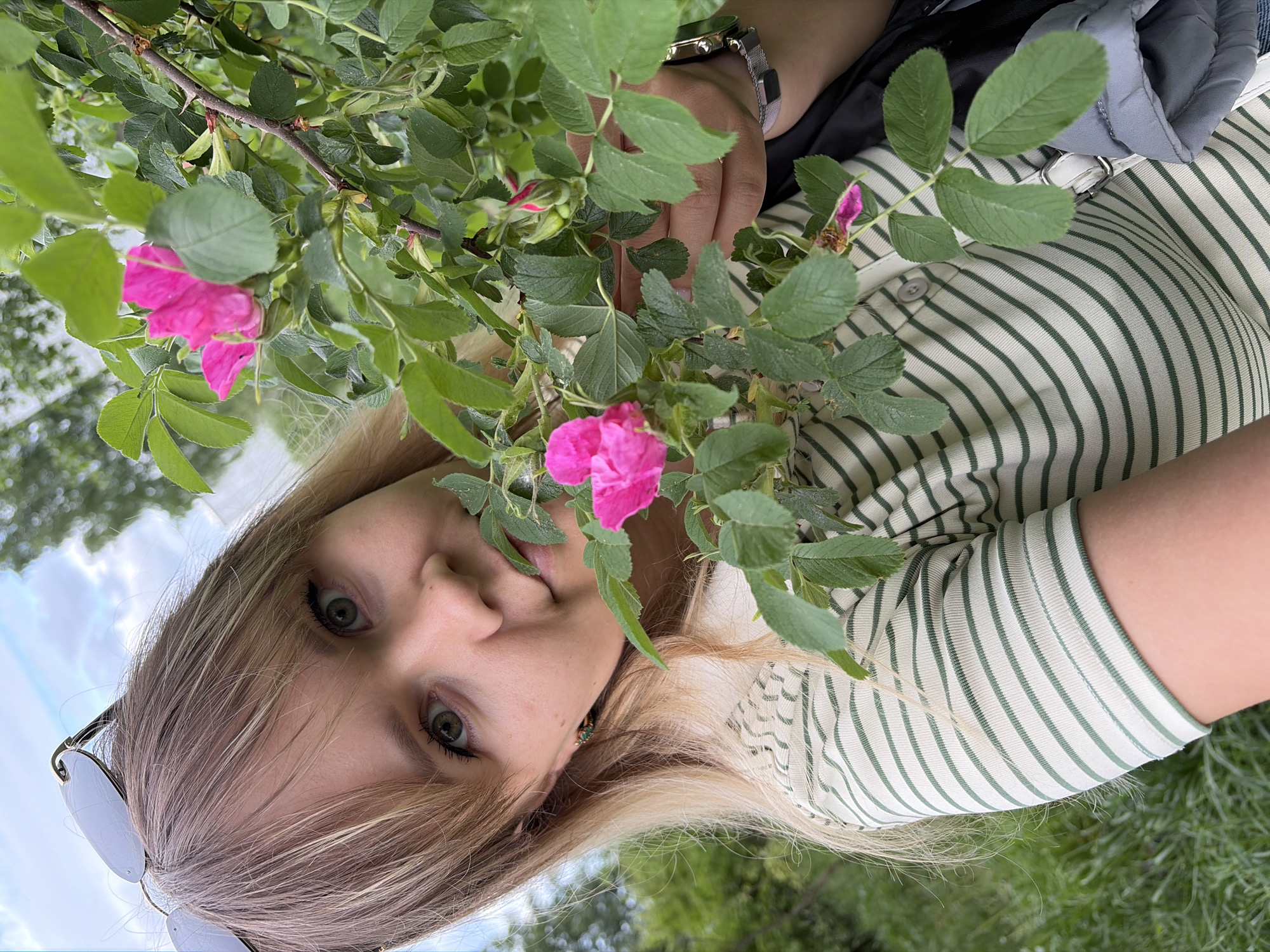 A young, blonde woman is positioned at an angle in an outdoor setting in Chaikovsky, Russia, with the horizon line running diagonally. Her face is tilted toward the viewer, and she wears sunglasses. The woman is adorned with a striped, long-sleeved shirt, and her hand, featuring a wristwatch, reaches into a cluster of greenery punctuated by pink blossoms, which are centrally located and partly obscured by the woman’s face. A backpack hangs over her shoulder. The background consists of lush greenery and patches of blue sky with clouds.