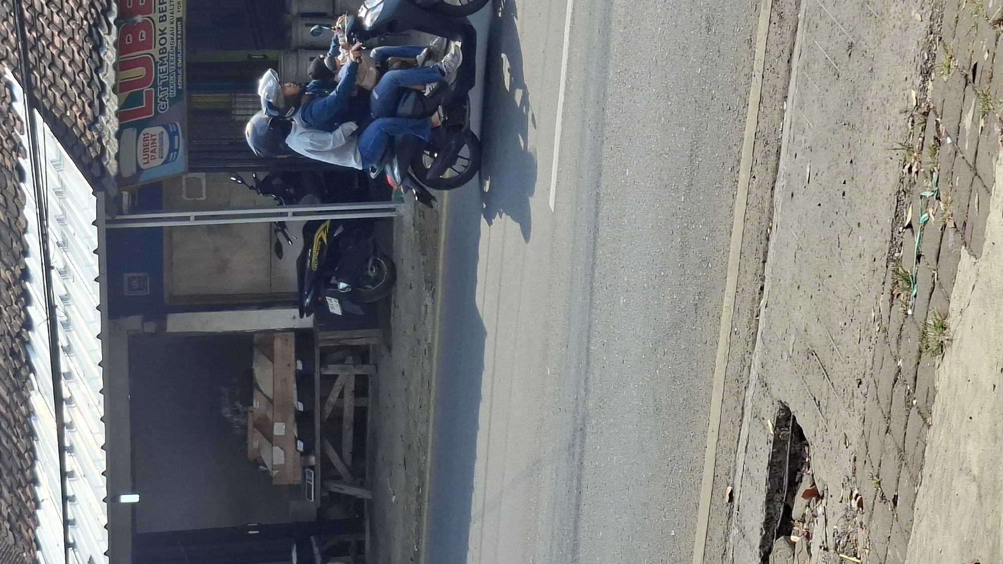 In Cilaku, Indonesia, two individuals are riding a scooter on a paved road. The person in the front is wearing a blue jacket and jeans, while the person behind them is wearing a light-colored long-sleeved shirt and jeans. Both are wearing helmets. The scooter is black and has a license plate that is partially visible. In the background, there is a building with a white roof and corrugated metal siding. A sign on the building reads "LUBERS PAINT". There are also several motorcycles parked in front of the building. The road in the foreground shows signs of wear and tear, with a broken section of pavement.