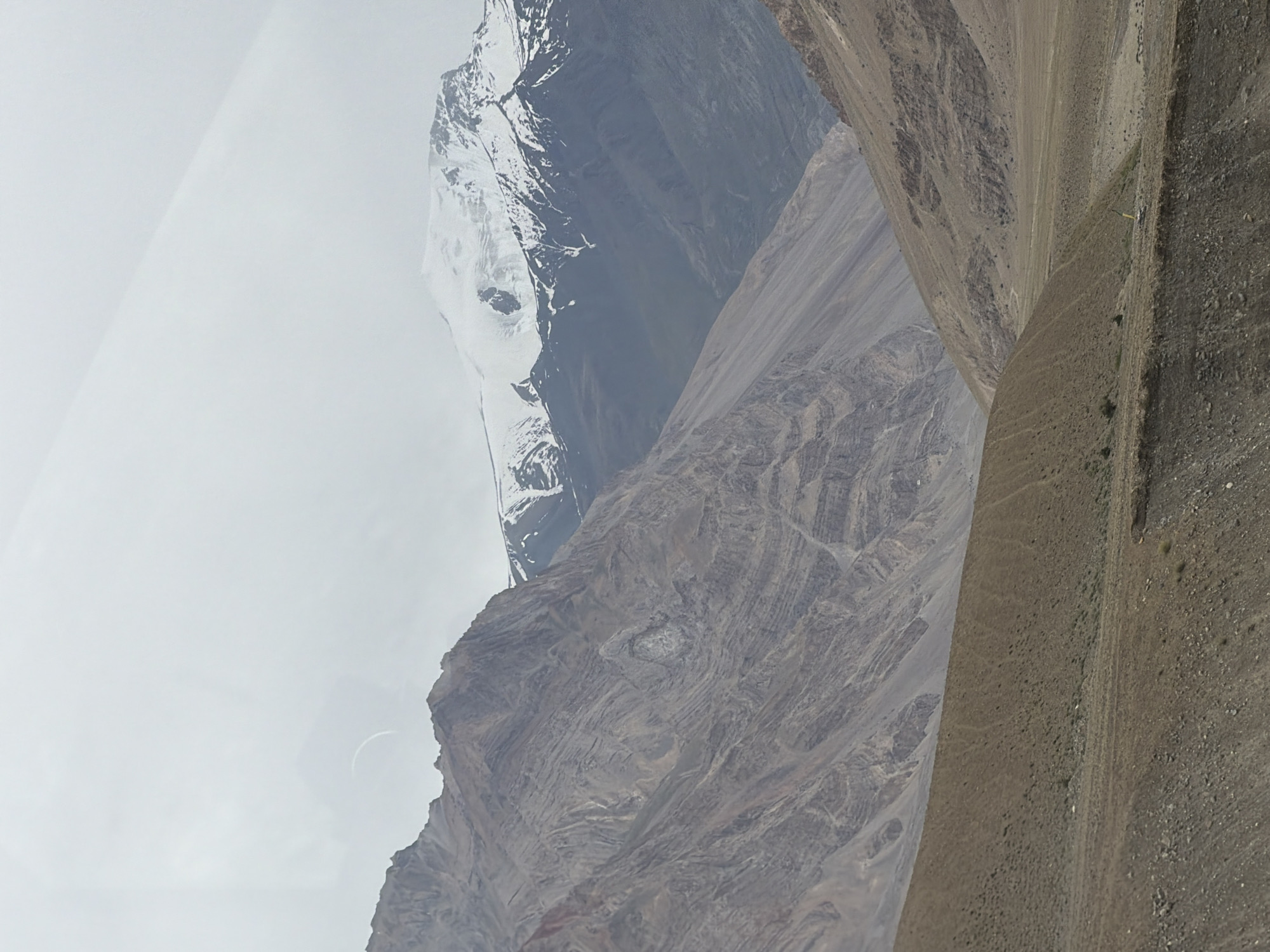 This is a scenic, eye-level, outdoor, vertical, full shot of a mountainous landscape in Zangla, India, under an overcast sky. In the distance, snow-capped mountains rise, with a bright, curved object, possibly a reflection or an artifact, visible in the sky above the left mountain range. The foreground shows a barren, brown, dirt slope with some sparse vegetation. The middle ground is dominated by rugged, layered mountains in shades of brown and gray, with visible striations suggesting geological formations. The background features more distant mountains, some with patches of snow. The overall scene is one of a vast, arid, and remote mountain environment.