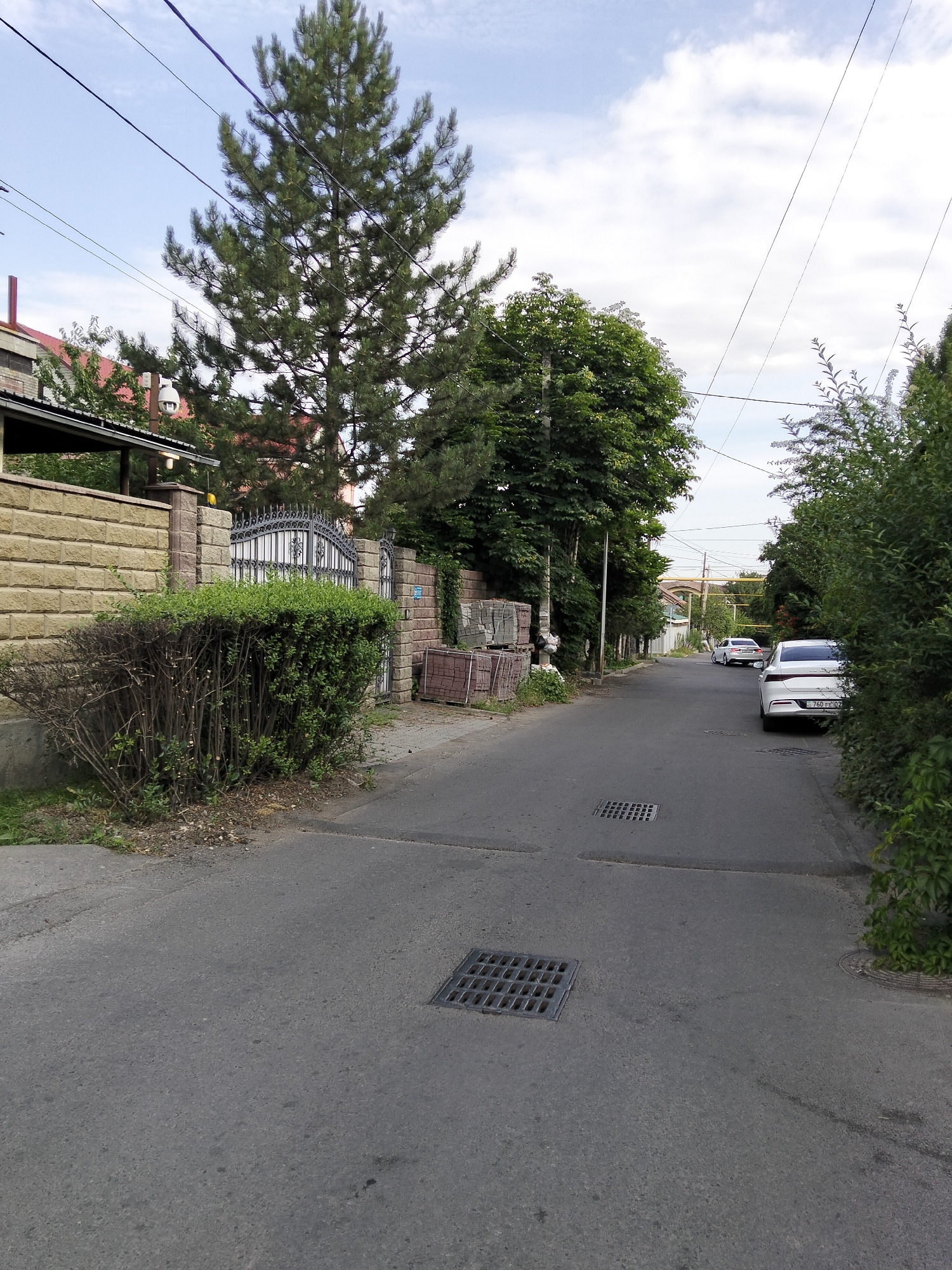 The image shows a street scene in Almaty, Kazakhstan. The street is paved and appears to be residential. There are buildings and fences lining the street, as well as trees and vegetation. Two cars are visible on the street, and there are also manhole covers. The sky is cloudy.