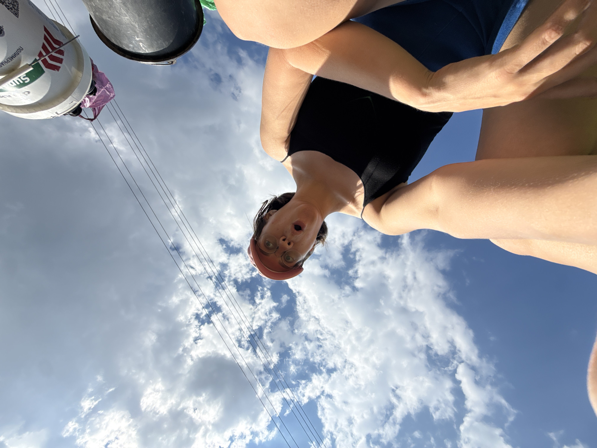 The image shows a low-angle, wide-shot perspective of a woman with a pink baseball cap and glasses looking upwards with a surprised expression. She is wearing a black tank top and blue shorts. Her arms are visible, with one hand resting on her leg and the other reaching towards the camera. In the background, there is a blue sky with white clouds, and power lines stretch across the frame. Two buckets, one white and one black, are visible hanging from the power lines. The white bucket has some red and green markings and a pink plastic bag is tied to it. The scene appears to be outdoors, possibly in an urban or suburban area, given the presence of power lines and buckets. There is no information in the image to indicate the specific location in the city of Dokshitsy, Russia.