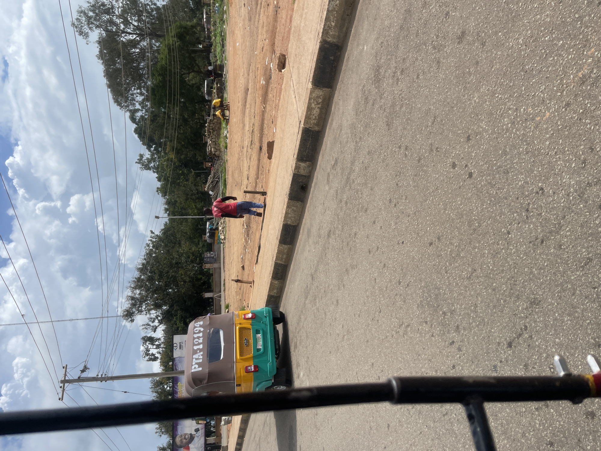 In Bar Jirgi Summa, Nigeria, a tricycle, painted green, yellow, and brown, with the license plate "PTA 12194" visible, is parked on the right side of a paved road. The tricycle's rear is facing the camera. The road has a concrete curb with alternating black and white stripes. A person in a red shirt and blue jeans walks away from the camera along the sidewalk. In the background, there is an undeveloped lot with dirt and scattered debris. Several trees are visible in the distance, and a power pole with numerous wires stands on the left side of the road. The sky is partly cloudy with large white cumulus clouds.