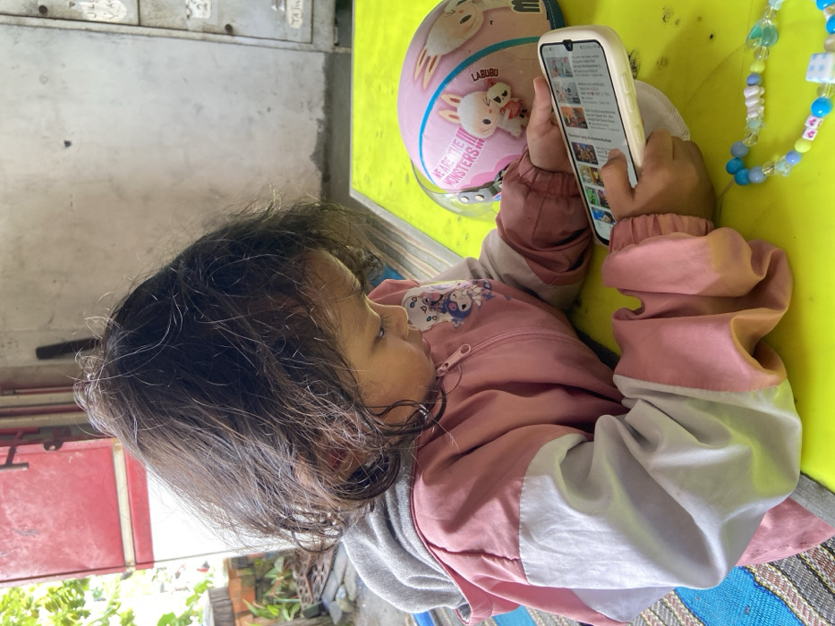 A young child is absorbed in watching a smartphone. She is wearing a pink jacket with white accents and has shoulder-length brown hair. The phone is held in her right hand, displaying a video platform, likely YouTube. Next to her is a pink helmet with cartoon characters and a colorful beaded bracelet. The background includes a light green surface and hints of an outdoor setting, suggesting she's in a casual environment.