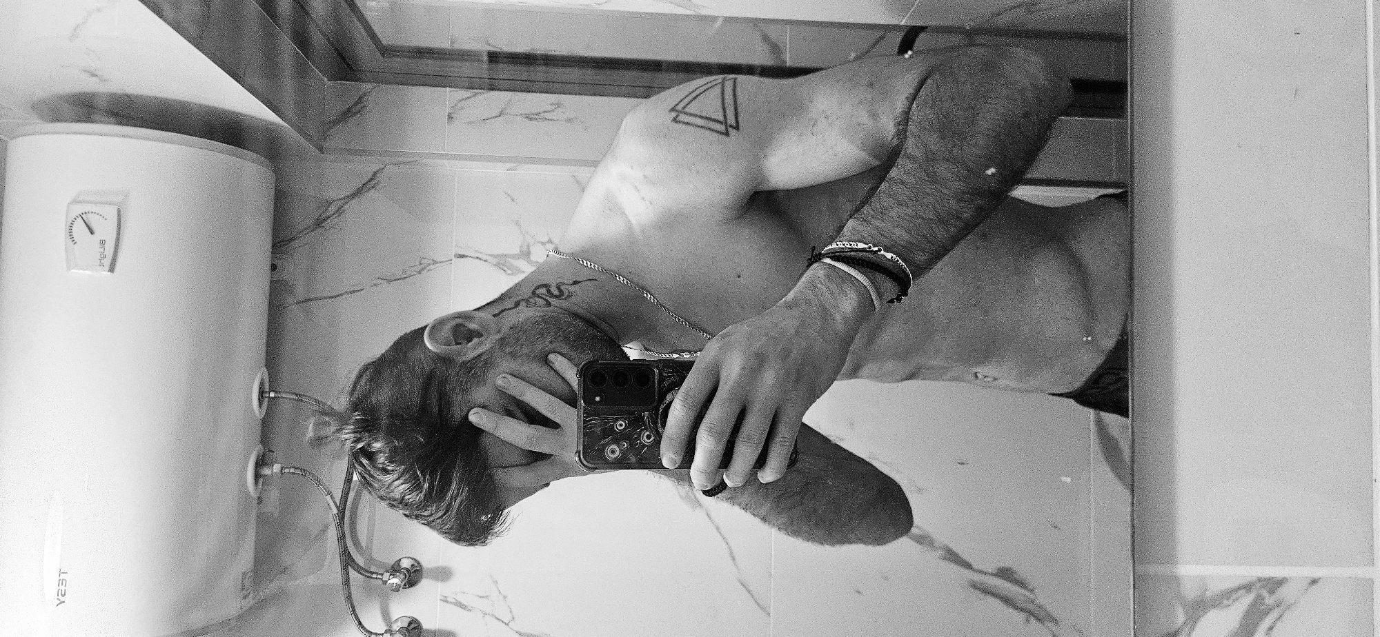 A black and white, inverted mirror selfie in a bathroom. A muscular man with tattoos on his arm, shoulder and neck holds a cell phone in front of his face. The backdrop includes marble walls and a water heater tank mounted on the wall. The image's location in Plovdiv, Bulgaria cannot be confirmed.