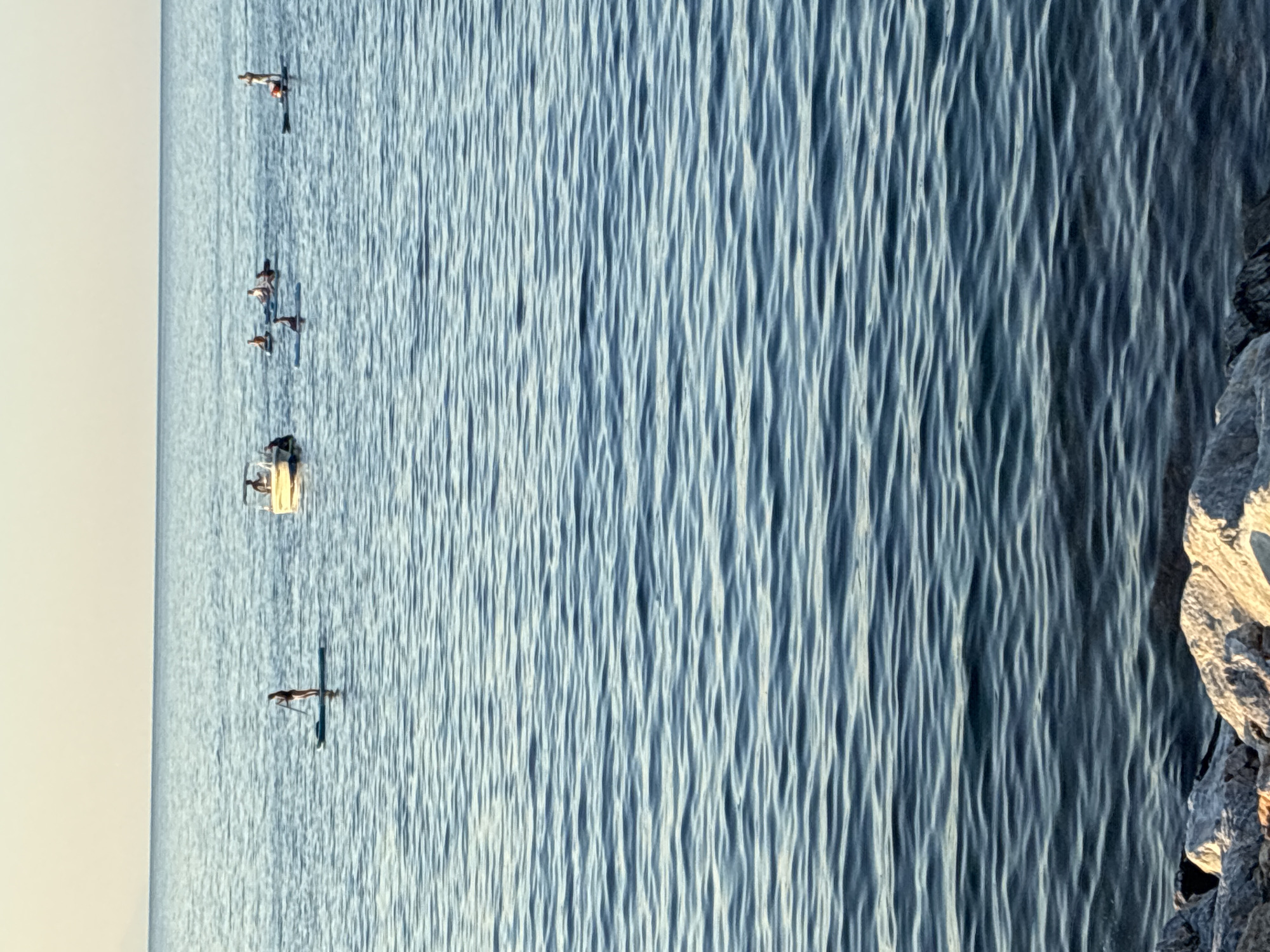 The image depicts a serene coastal scene with the Mediterranean Sea in Alanya, Turkey, under a clear, pale sky. Several individuals are engaged in water activities. In the middle ground, a woman stands on a paddleboard, holding a paddle. To her right, a small white boat with two occupants is visible. Further to the right, a group of people are also on paddleboards, with some paddling and others in seated positions. The water is calm, with gentle ripples reflecting the soft light. In the foreground, rocks and pebbles line the shore, suggesting a natural beach environment. The overall impression is one of leisure and recreation on a peaceful day.