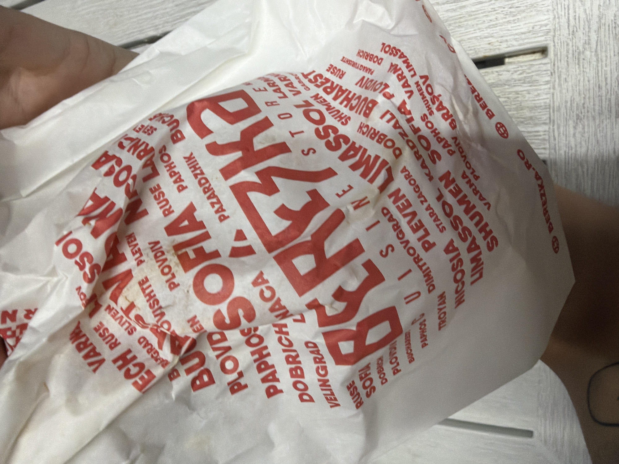 The image shows a close-up of a white plastic shopping bag with red text. The bag is crumpled and being held by a person whose hand and arm are partially visible on the right side of the image. The text on the bag appears to be a list of city names, many of which are likely in Bulgaria and Cyprus, given the presence of "Sofia, Bulgaria" in the text. Some of the visible city names include VARNA, RUSE, SOFIA, PLOVDIV, LIMASSOL, BUCHAREST, NICOSIA, and PAPHOS. The bag has a large central logo or brand name printed in red, which appears to be "BERE**KA". The overall impression is of a shopping bag from a retail chain with a presence in multiple cities.