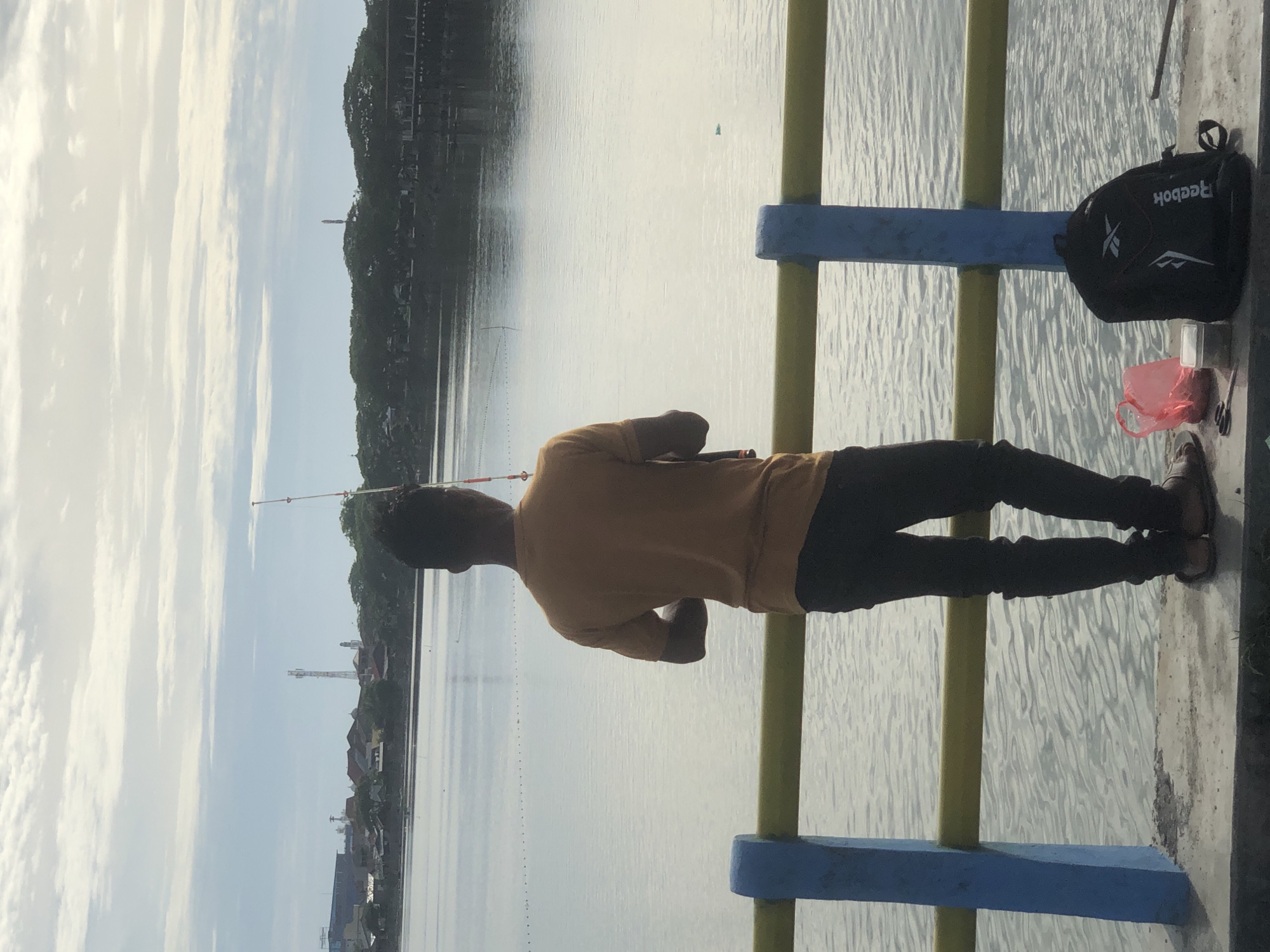 A person is fishing off a bridge railing over a body of water. The person is wearing a mustard-colored shirt and dark pants. A black Reebok backpack is next to him on the railing. There is a red plastic bag, a pair of scissors and what seems to be fishing tools next to the backpack. A fishing pole is visible extending out over the water. The scene appears to be located in Lhokseumawe, Indonesia, given the buildings in the distance and the general architecture.