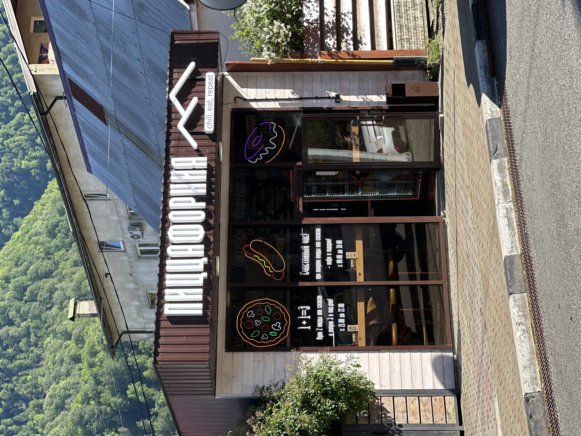 An exterior, slightly tilted view captures the storefront of a food establishment named "ПИЦЦАФОРНИЯ" (Pizzifornia) on a sunny day in Krasnaya Polyana, Russia. The building features a dark brown roof and lighter, possibly wooden, lower walls, with large glass windows and doors forming the main entrance. A prominent white sign on the facade displays the restaurant's name in Russian, with a smaller white sign below it in English that reads "chill, eat, repeat."
Through the glass, colorful neon-style signs illustrate a donut, a hot dog, and a pizza, indicating the menu offerings. Several promotional texts are displayed on the windows in Russian: "1+1=3 Купи 2 пиццы или сосиски и получи 3 в подарок! с 21:00 до 22:00" (Buy 2 pizzas or sausages and get 3 as a gift! from 9 PM to 10 PM) and "Счастливый час! при покупке пиццы или СОСИСКИ - кофе в подарок! с 09:00 до 10:00" (Happy hour! coffee as a gift with pizza or sausage purchase! from 9 AM to 10 AM). Other partially visible Russian text includes "СОБЕРИ СВС ПИЩА ФОРНИК)", "ХАЛАРЕНЬО" (jalapeño), "ОГУРЕЦ МАРИНОВАННЫЙ" (pickled cucumber), and "топинг" (topping), likely describing menu items or ingredients.
Lush green bushes are planted along the base of the building, and flowering plants are visible on an upper balcony. To the right, a paved sidewalk with a curb borders an asphalt road. The background reveals a steep, densely forested mountain slope, characteristic of the region. The strong sunlight and clear shadows indicate it is daytime. An air conditioning unit is mounted on the building's upper level. No specific activities or individuals are clearly identifiable, but the establishment appears to be open for business.