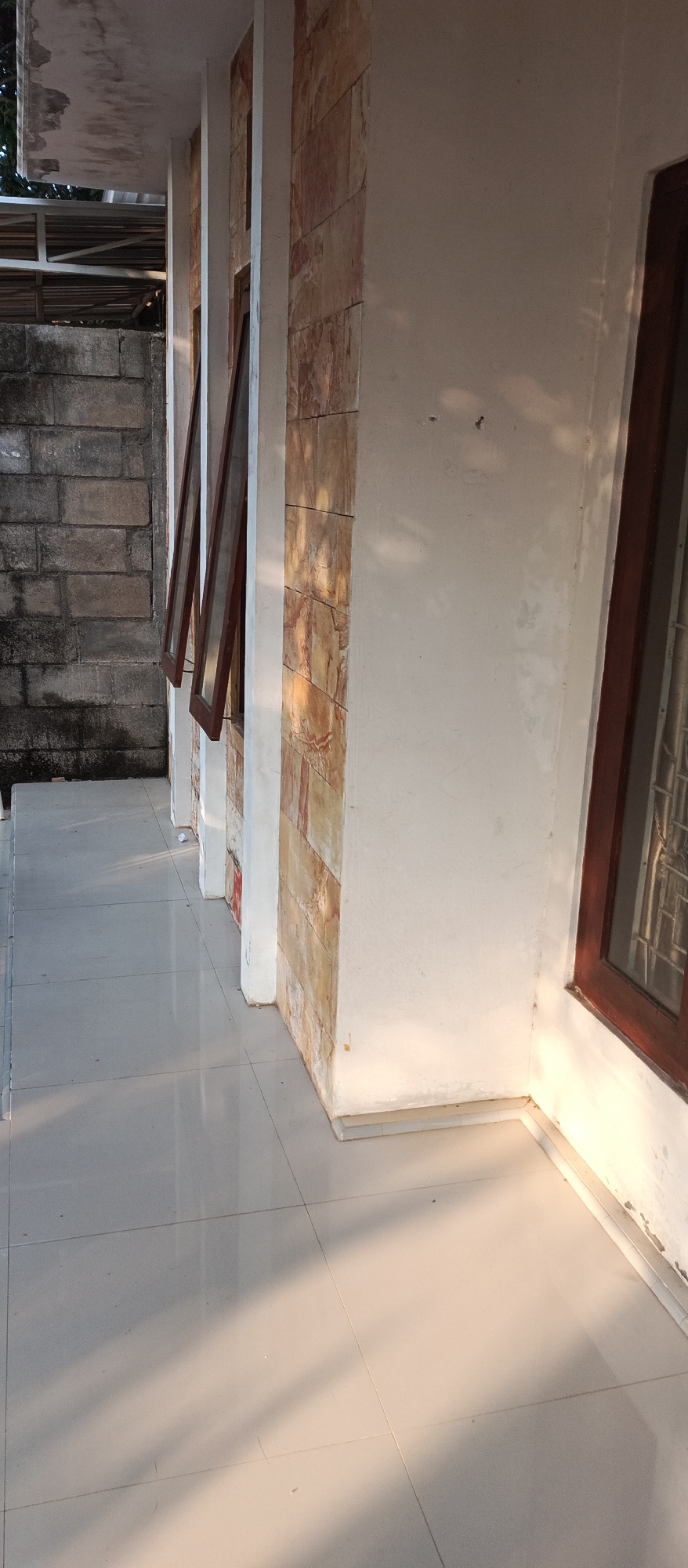 The image shows an exterior view of a building in Jatisari, Indonesia. The foreground is dominated by a tiled floor, with light reflecting off its smooth surface. Several rectangular columns, clad in a textured stone material of earthy tones, are visible. Between these columns, partially open windows with wooden frames are set into the wall. The wall itself is a light, off-white color. To the left of the building, a rough, grey brick wall forms a boundary. The scene is illuminated by warm, low-angled sunlight, casting distinct shadows across the floor and highlighting the textures of the stone and walls. The overall impression is one of an outdoor walkway or porch area adjacent to a residential or commercial building.