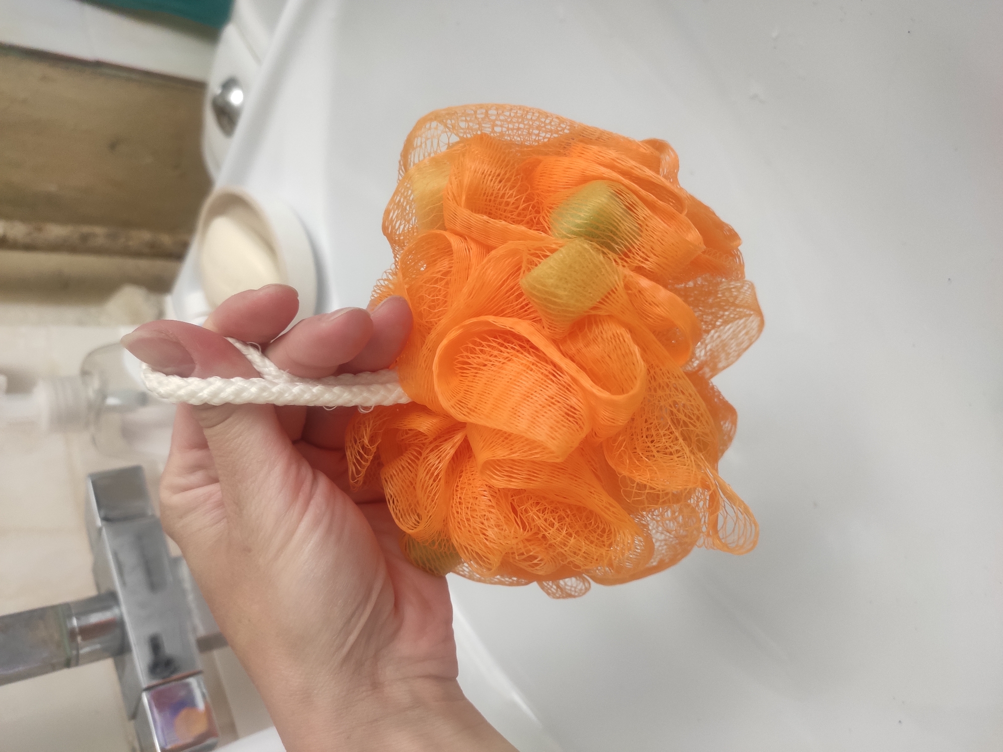 A hand holds an orange loofah in a bathroom setting. A white sink, faucet, and soap dispenser are visible in the background. The scene appears to be indoors. There is no evidence in the image to determine the location in Sterlitamak, Russia.