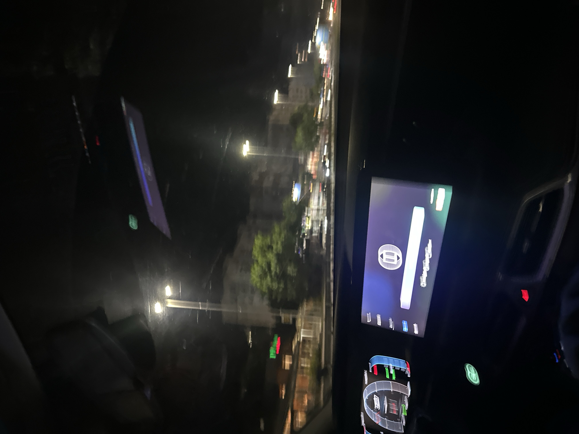 The image is a low-light, first-person perspective from inside a car at night, likely in the city of Shumen, Bulgaria. The view is through the windshield, showing a street scene with buildings, trees, and streetlights. The car's dashboard is visible in the foreground, displaying a digital instrument cluster on the left and a central infotainment screen on the right. The infotainment screen shows a paused media player interface. The overall impression is of a night drive through an urban area, with reflections on the windshield contributing to the atmospheric quality of the image.