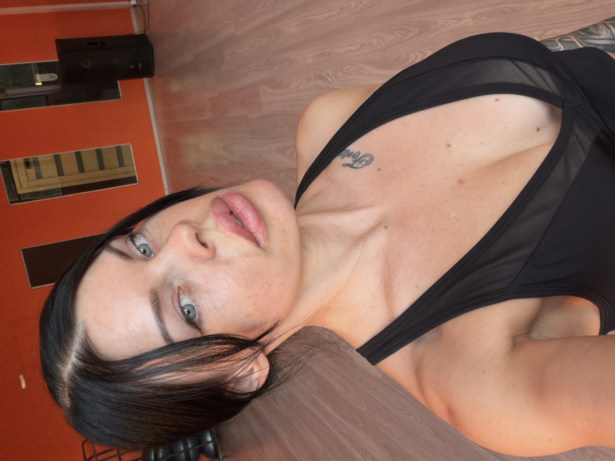 A woman with dark hair and light blue eyes, wearing a black athletic top, takes a selfie in a room with orange walls and a wooden floor. The room appears to be a fitness studio or gym, with weights racks visible in the background and a large speaker unit. The woman has a tattoo on her collarbone. The specific location is not provided in the image or surrounding context.