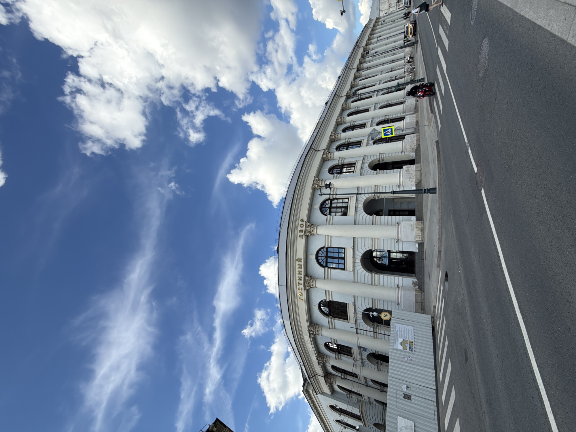 The media file depicts a wide, curved building with classical architecture, likely a historical landmark or public institution. The building features a colonnade with large white columns and arched windows. The text "ГОСТИНЫЙ ДВОР" is visible in gold lettering above a section of the building, indicating it is the Gostiny Dvor, a historic trade arcade in Moscow, Russia.
The scene is an outdoor street view. The building's facade is a prominent subject, stretching along the right side of the frame. In the foreground, a paved road with a pedestrian crossing is visible. A motorcycle is parked on the street near the crossing, and a person is walking across it in the background. Another person is visible further down the street, near a yellow car.
The sky above is a bright blue with scattered white clouds, suggesting a clear and sunny day, likely daytime. A yellow traffic sign with a pedestrian symbol is mounted on a pole in front of the building. A temporary construction barrier or hoarding is present on the sidewalk in front of a section of the building, with some posters or notices attached to it. The overall impression is of a bustling urban environment with a mix of historical architecture and modern street life.