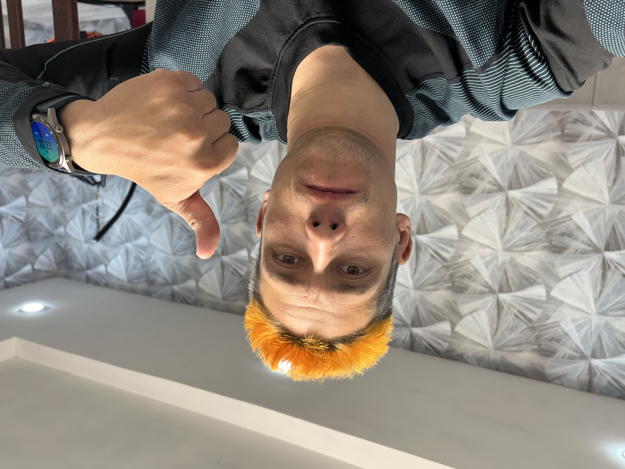 The image, rotated 90 degrees counter-clockwise, depicts a man from the chest up, looking directly towards the viewer. He appears to be looking upwards, as if at a camera positioned above him or towards a ceiling. He has short dark hair with a distinctive bright orange, short fringe or bang across his forehead. He sports a light stubble beard and has a neutral to slightly serious expression.
He is wearing a long-sleeved shirt that is black on the main body and collar, with black and blue/teal patterned fabric on the sleeves and shoulders, featuring both small geometric dots and horizontal stripes. On his left wrist, he wears a metallic grey smartwatch with a dark strap, the screen of which clearly displays "22:22" and other digital information, indicating the time is 10:22 PM. His left hand is raised, making a clear "thumbs down" gesture.
The background behind the man features a textured wall with a repeating geometric pattern of white and grey fan-like or starburst shapes. Below the wall, a plain white ceiling with a single illuminated recessed spotlight is visible. The setting is an indoor environment, likely in Chaikovsky, Russia, as per the provided context. The gesture suggests disapproval or a negative sentiment.
