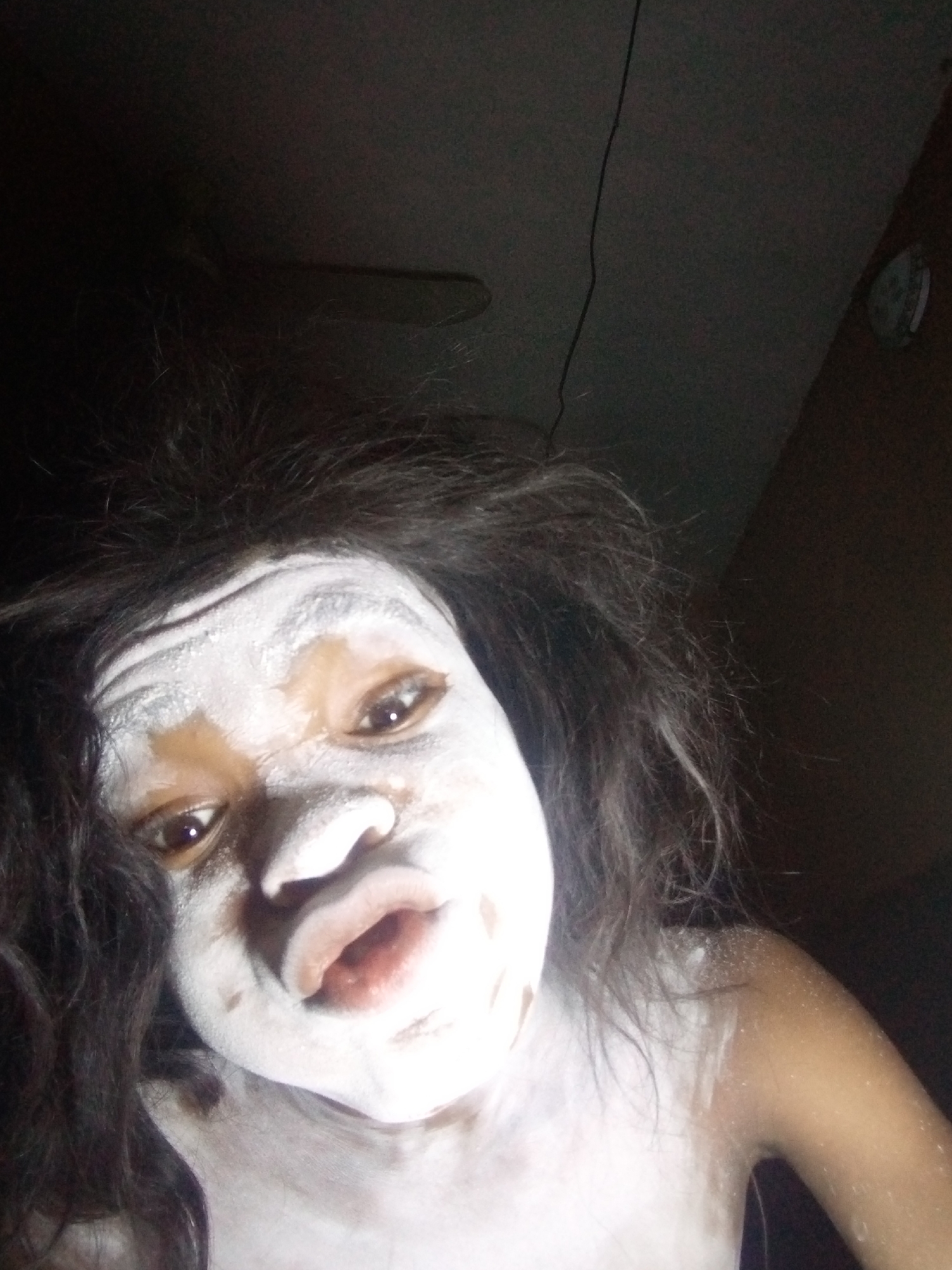 This image features a close-up of a person, likely a child or young adult, in a dimly lit indoor environment in Onitsha, Nigeria. The individual's face, neck, and shoulders are extensively covered in a white powdery substance, resembling chalk or white clay, which creates a striking contrast against their natural dark skin tone and dark, disheveled hair.
The subject's eyes are wide open, gazing directly towards the camera, and their mouth is slightly agape in a puckered or O-shaped expression. The lighting is harsh and direct, likely from a flash, which brightly illuminates the white substance on their skin while leaving the background largely in shadow.
In the blurred background, faint details suggest an ordinary room: a ceiling fan is partially visible at the top left, a dark electrical cord runs vertically on the left side, and a circular object, possibly a clock, can be seen on the upper right wall.
The application of white clay or similar substances is culturally significant in some Nigerian traditions, often used in rituals, masquerades, or as body adornment for various ceremonies or celebrations. The person's pose and the distinctive appearance suggest either participation in such a cultural event, a theatrical portrayal, or a playful act. No visible text is present in the image.